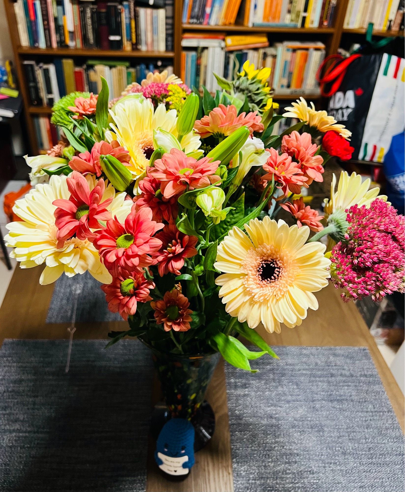 Photo of a vase of pink and yellow flowers sitting in a table in front of an overflowing bookcase.