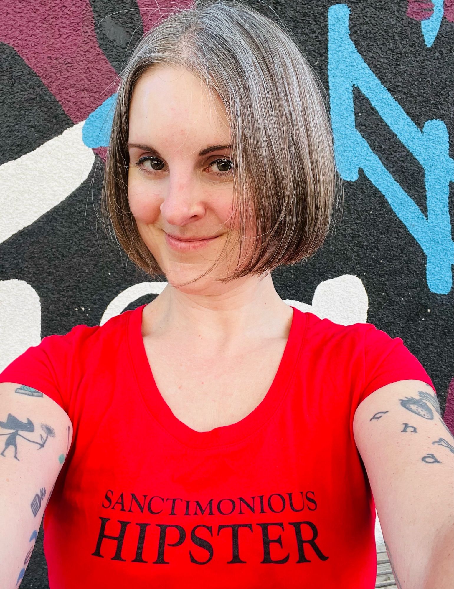 Selfie of Jen wearing a red shirt that says sanctimonious hipster in black letters standing in front of a colourful mural.