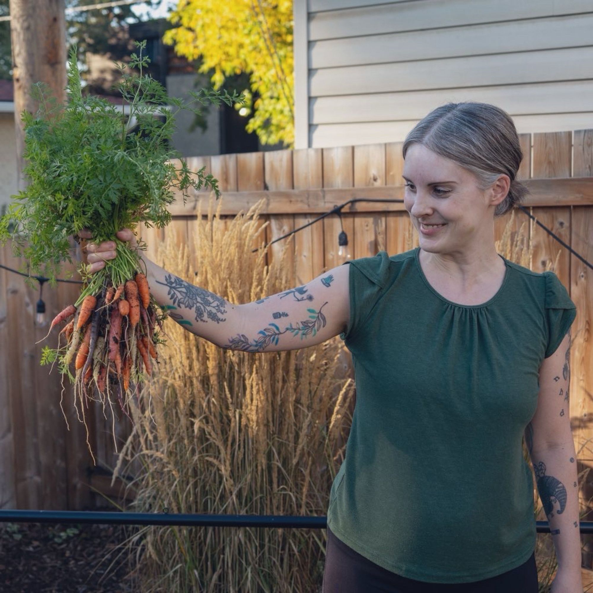 Photo of Jen in the backyard wearing a green T-shirt and black yoga pants holding a bunch of freshly picked garden carrots and smiling at them affectionately,