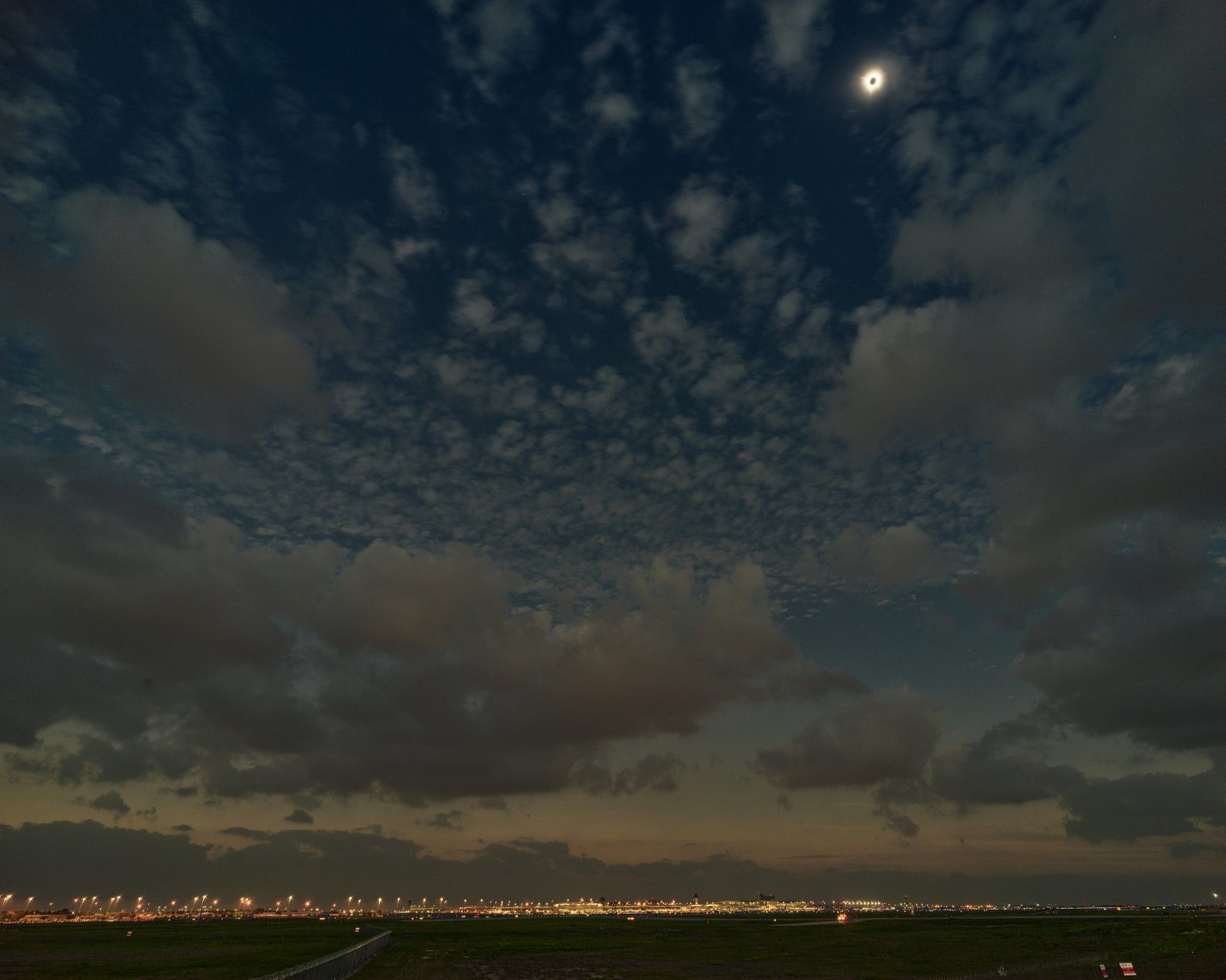 Solar eclipse at totality. A small, bright corona with a dark center visible at upper right. Most of the frame is a dark sky filled with various clouds, with a dark airfield visible at bottom. Airport terminal lights visible in background.