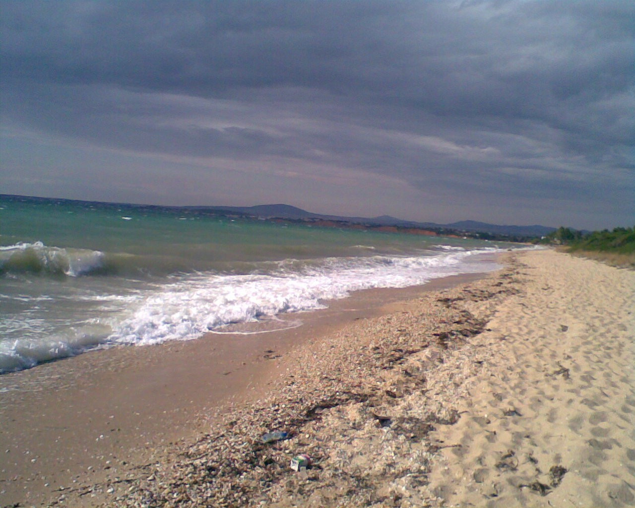 Sandstrand, dunkler Sand, schäumende Wellen, sehr dunkle Wolken, rechts etwas Grün. Im Hintergrund: niedrige Berge.