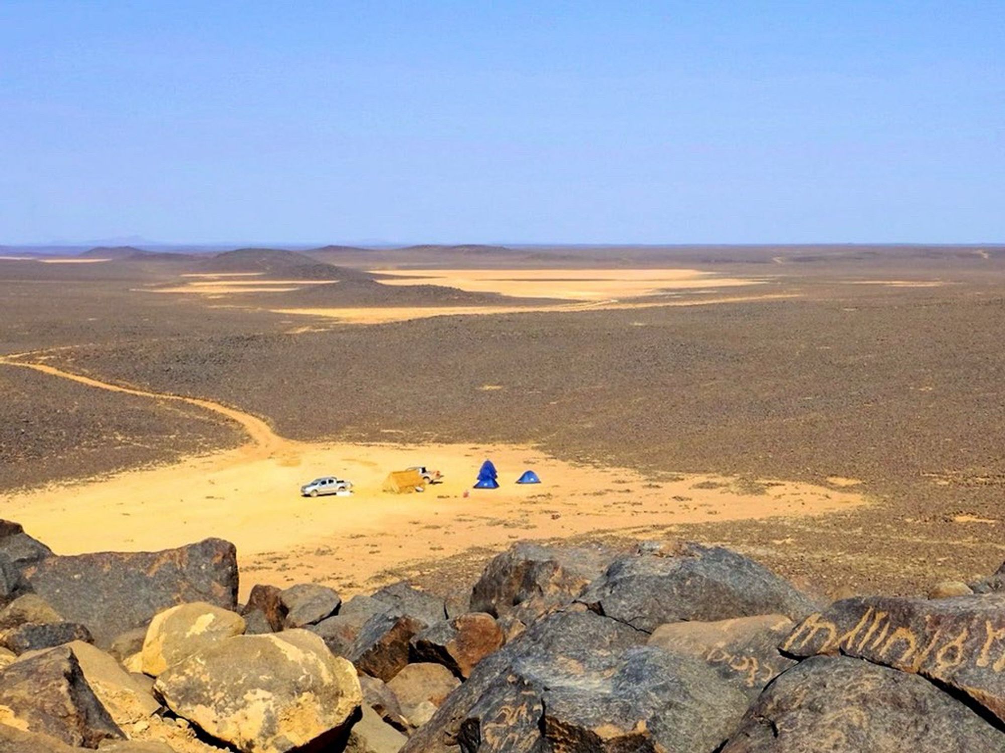 Photo of a dustpan in an otherwise rocky basalt desert. Taken from a higher point we are looking down at a large yellow-brown and four smaller blue tents making up a camp in the middle of the dustpan. A small pick-up truck is parking next to them. In the background another dustpan can be seen under the light blue sky in the distance.