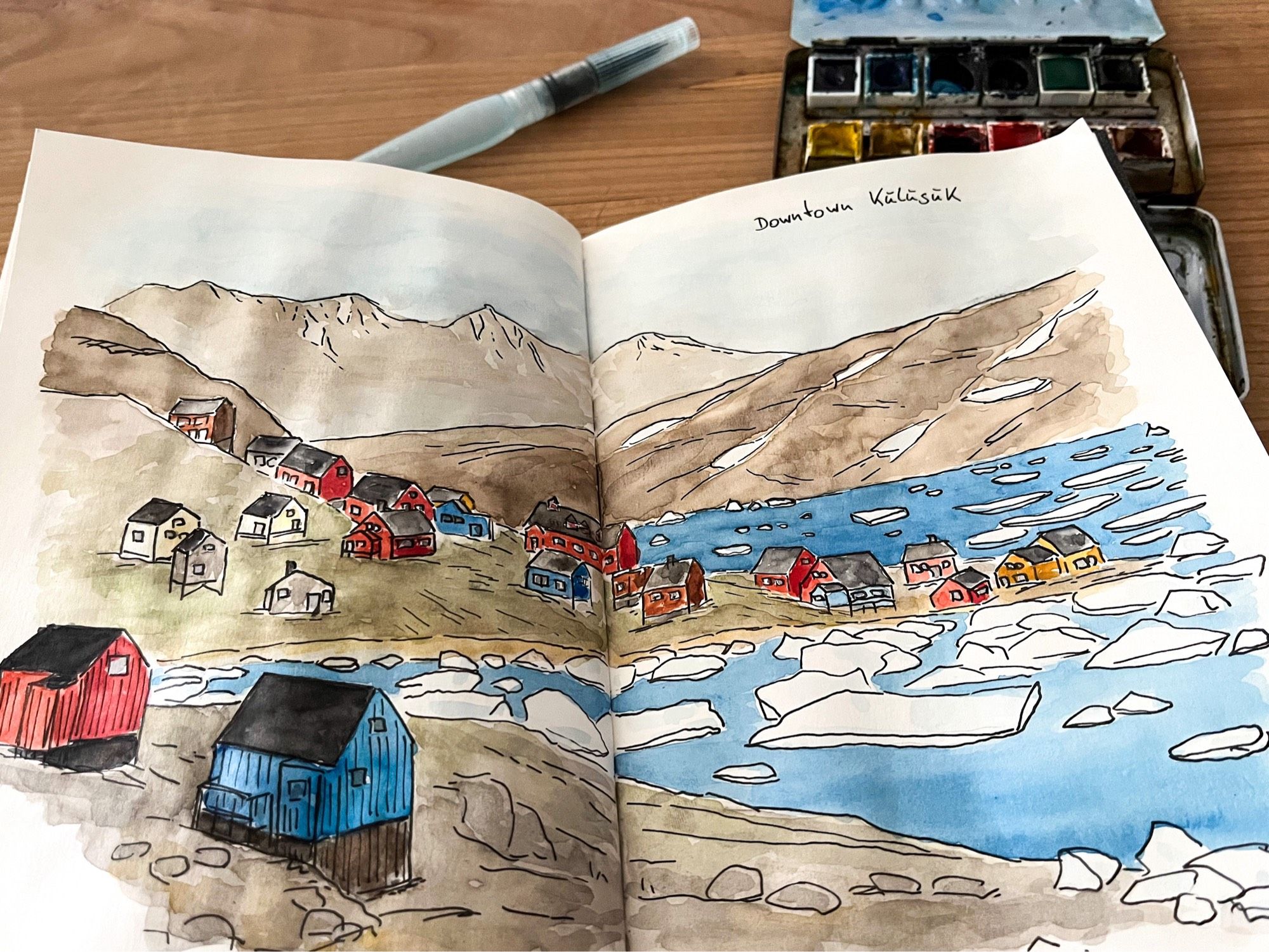 Foto of an open sketchbook showing about a dozen small colourful houses alongside an ice-filled Arctic fjord.