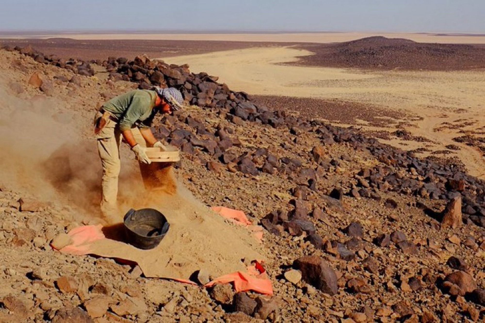 Photo of a man standing on the basalt-covered flanks of a slope, sifting bright soil, dust all around him. He is wearing a green long-sleeve shirt, khaki pants, and a headscarf. The soil he is sifting is accumulated on an orange tarp, an empty rubber basket can be seen at his feet. The background is a desert panorama, dust and basalt and a bright sky.