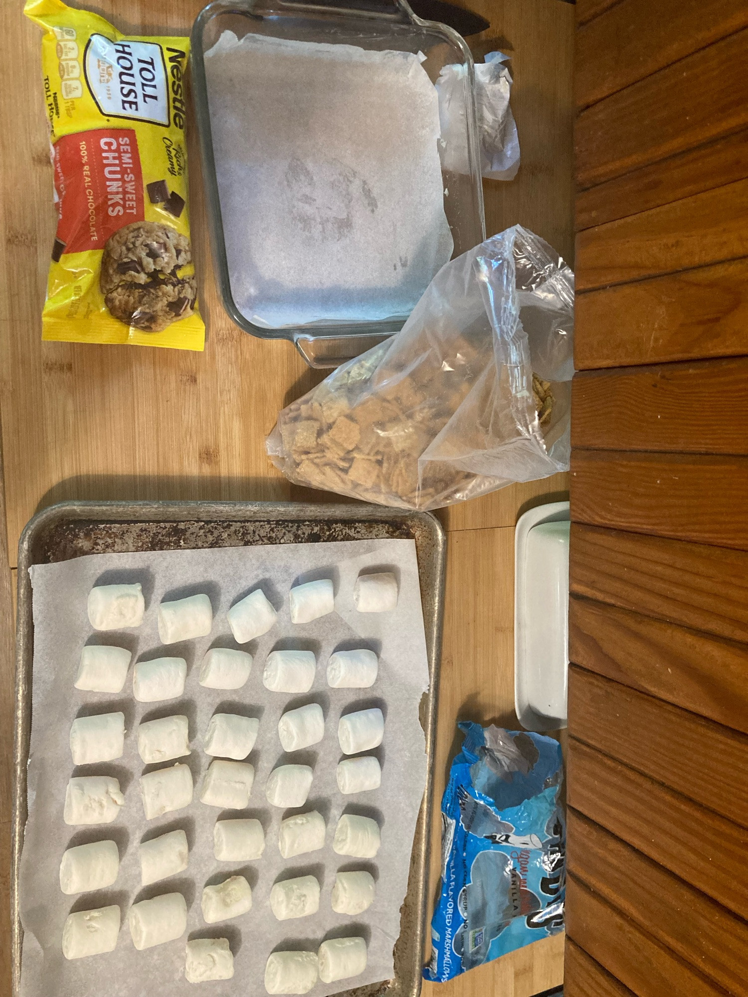 Kitchen still life of a bag of chocolate chunks, square cake pan lined with parchment, bag of graham cracker cereal, a cookie sheet pan with marshmallows, empty bag of marshmallows, butter dish.