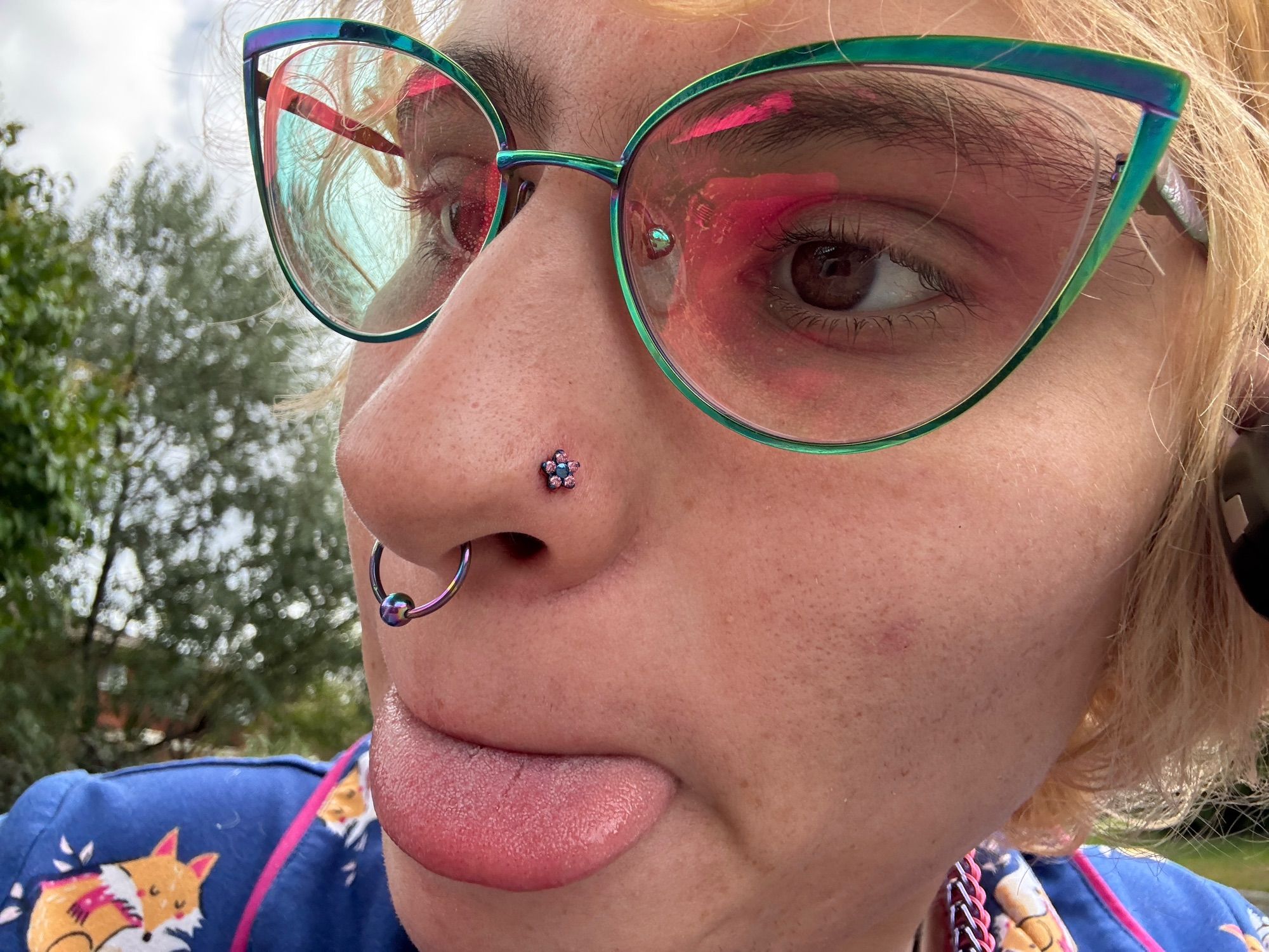 A close up of our right nostril piercing it is a pink flower with a blue inside flower. we have our rainbow septum piercing showing. We have our rainbow cat eye glasses on and we’re blepping.