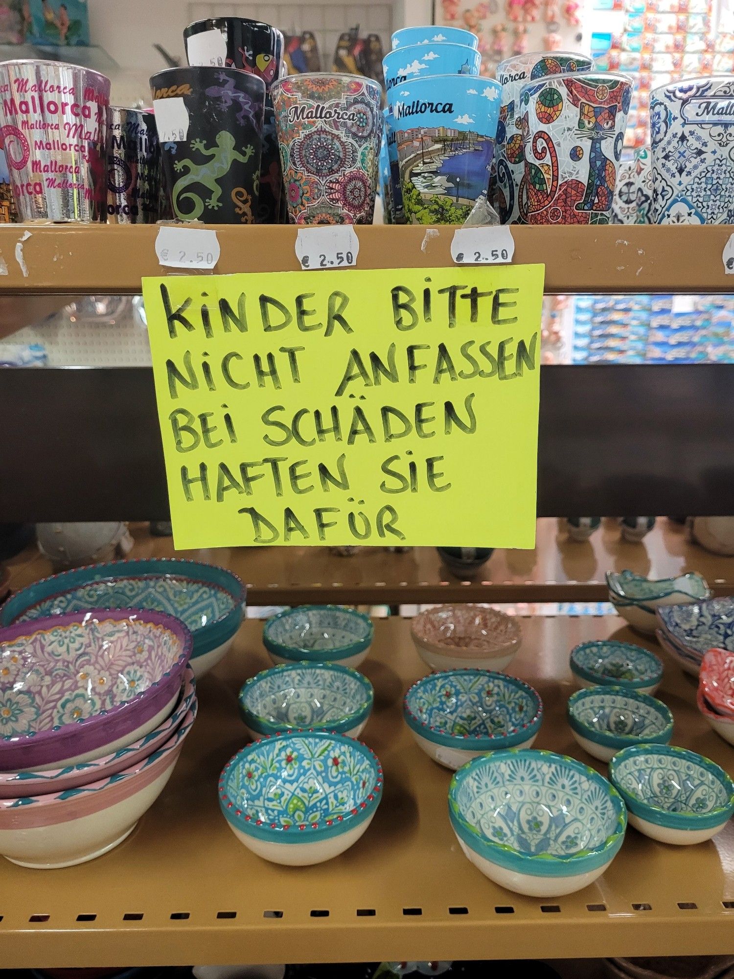 Ein gelbes, habdgeschriebenes Schild "Kinder bitte nicht anfassen. Bei Schäden haften sie dafür." In den Regalen daneben sind einige Becher, Schüsseln etc zu sehen.