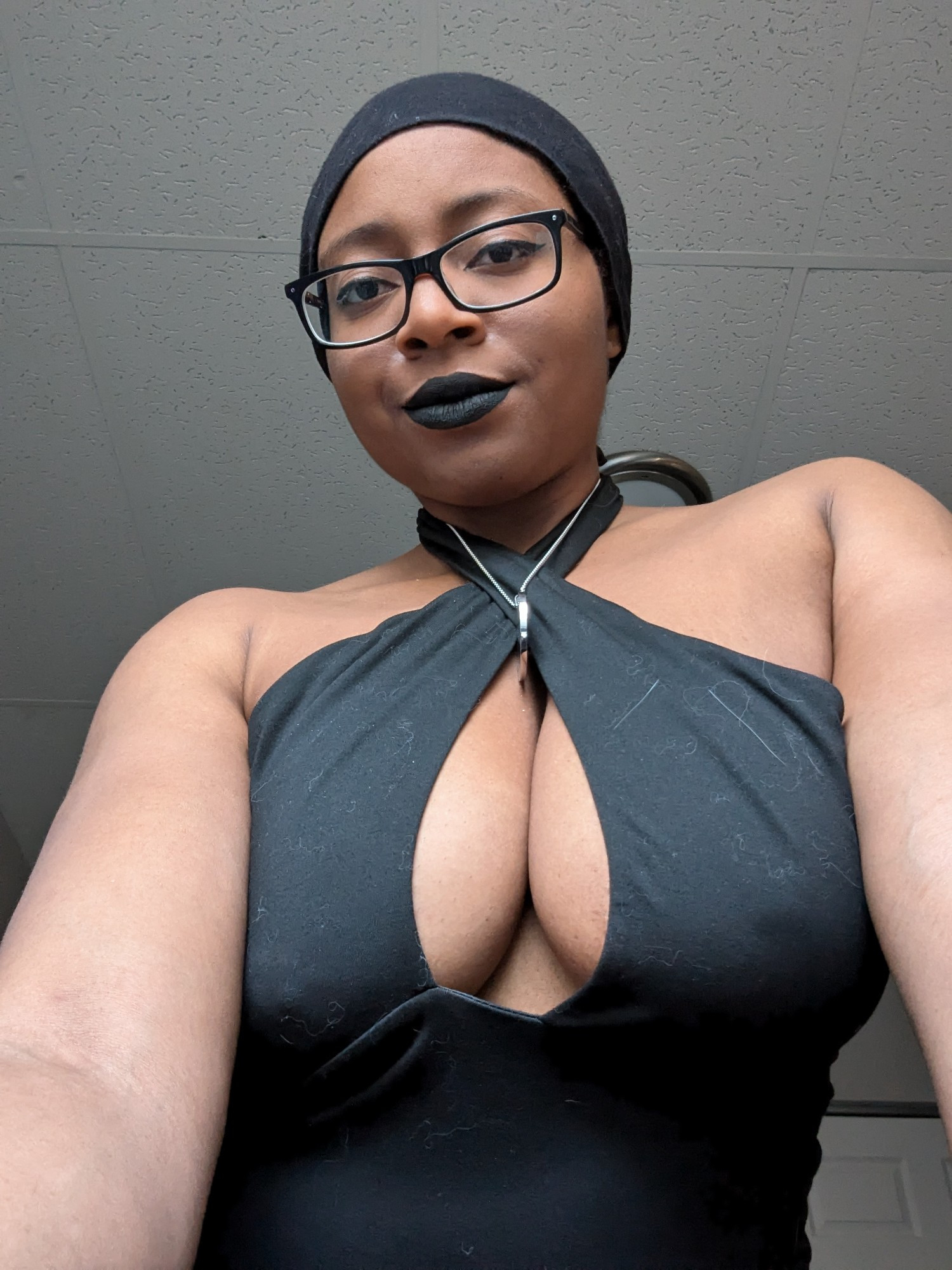 Waist up selfie of gothic Black American woman wearing black rimmed glasses, black eyeliner, black lipstick, and a black crisscross top 