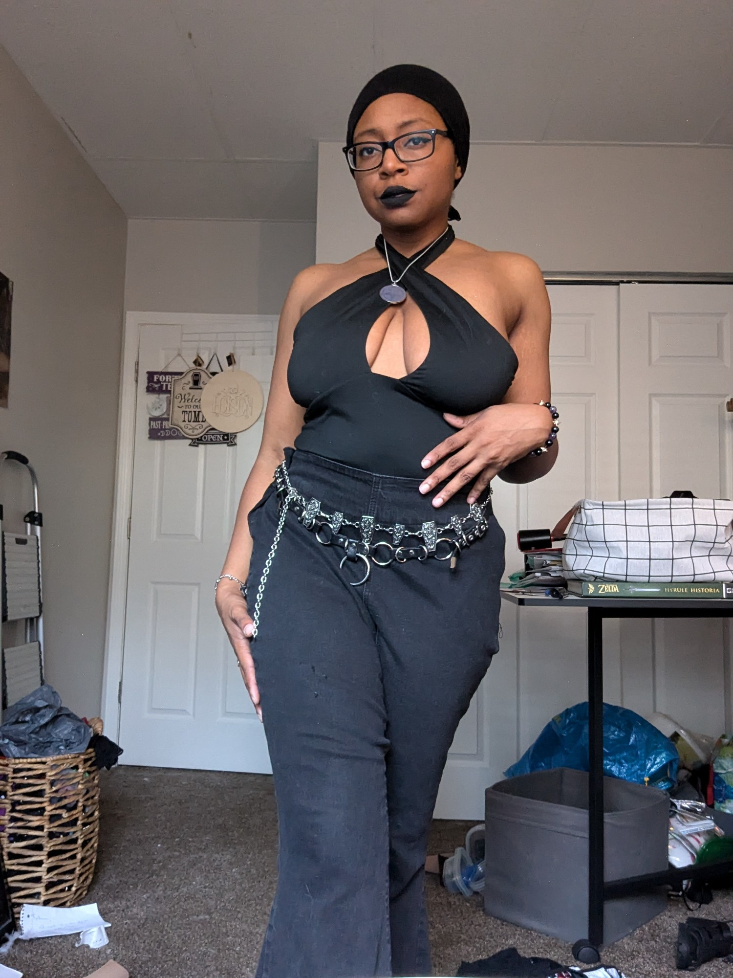 Photo of gothic Black American woman wearing black rimmed glasses, black eyeliner, black lipstick, a black crisscross top, black jeans and belts