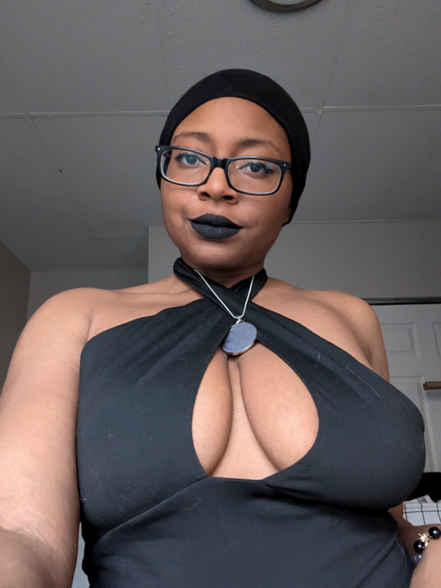 Waist up selfie of gothic Black American woman wearing black rimmed glasses, black eyeliner, black lipstick, and a black crisscross top 