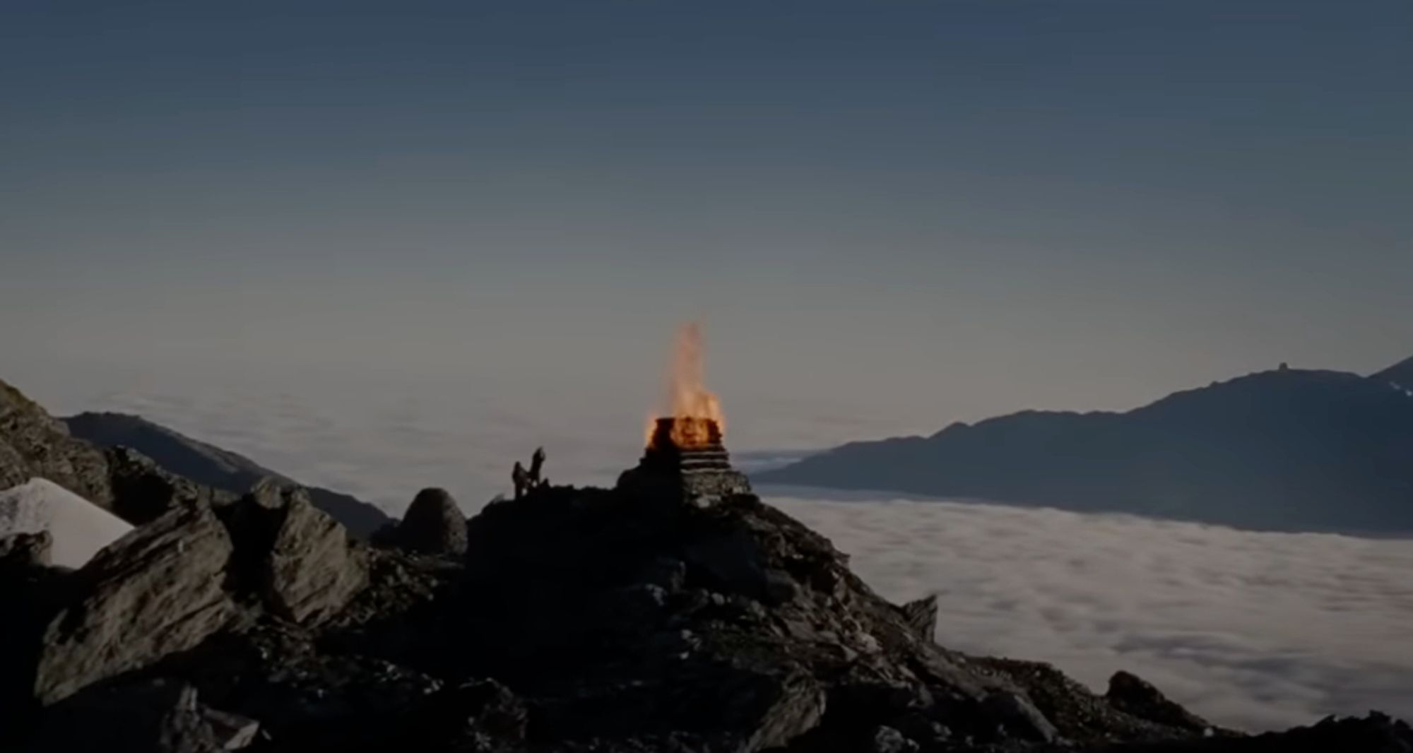 A screenshot from The Lord of the Rings: The Two Towers. A small figure can be seen next to a fire beacon on top of a mountain that sits above the clouds in a great mountain range.