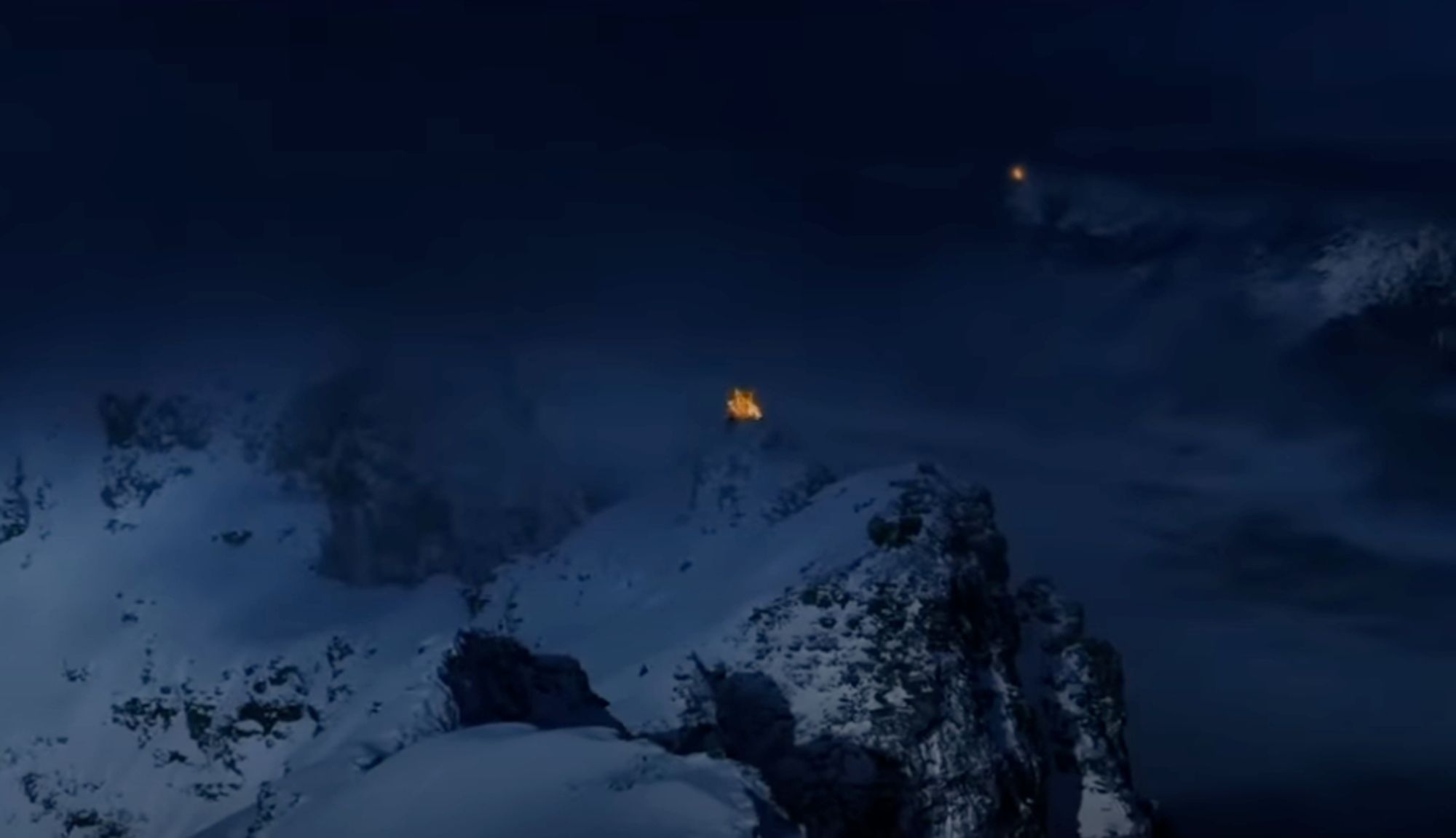 A screenshot from The Lord of the Rings: The Two Towers. It's a blue-hued night shot of a snowy mountain range with another far in the distance. Both peaks have large fire beacons on top.