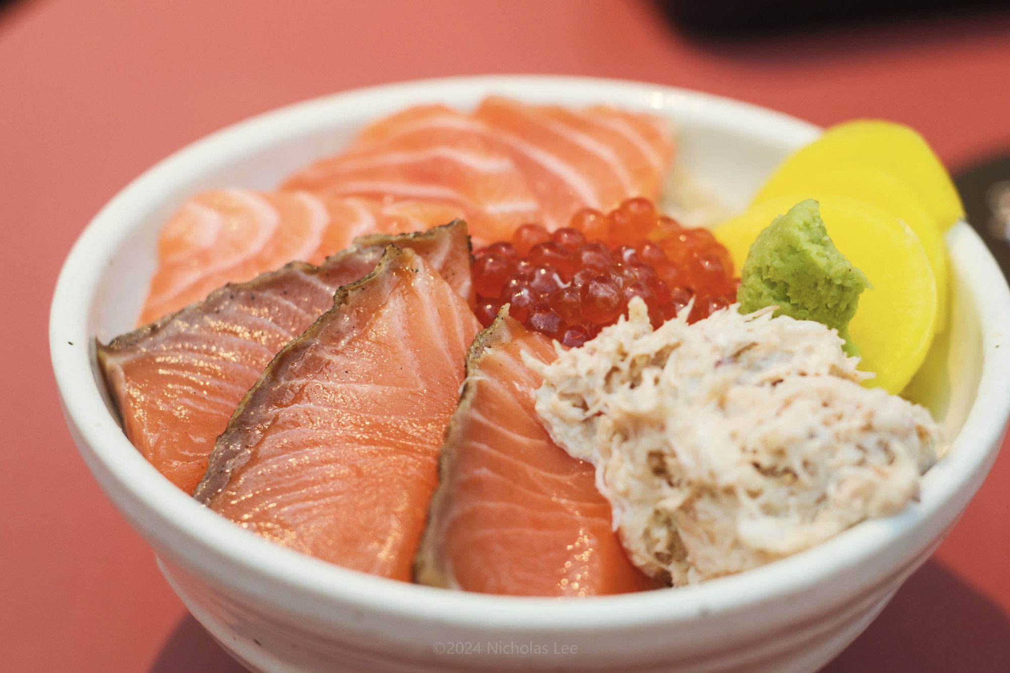 Close up of a bowl of sliced raw salmon, aburi salmon, ikura and snowcrab meat on sushi rice.