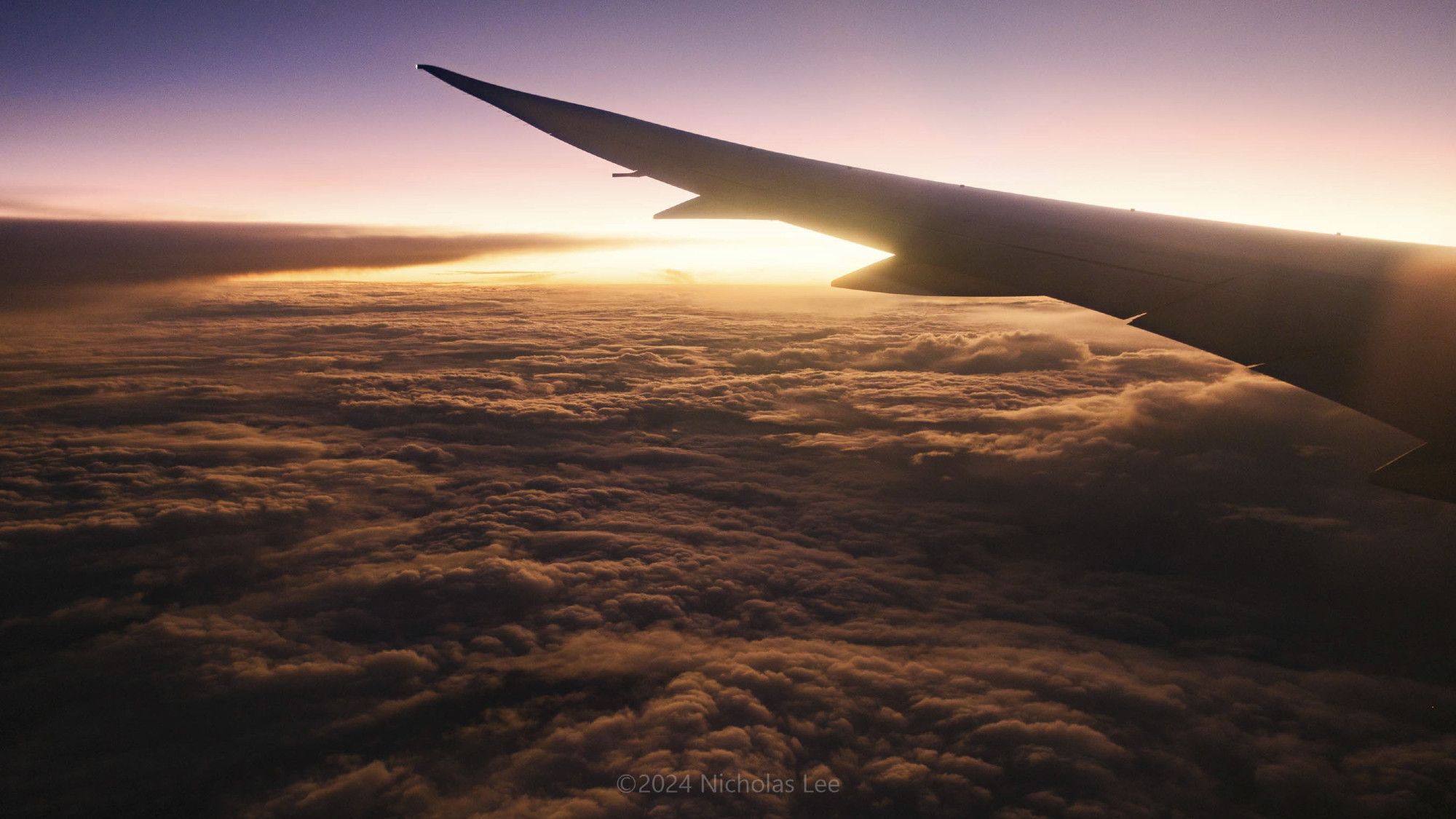 Photograph of a warm sunrise from a plane window. The sunlight is streaming warm light through the planes wing and casting long shadows on the bed of clouds below.