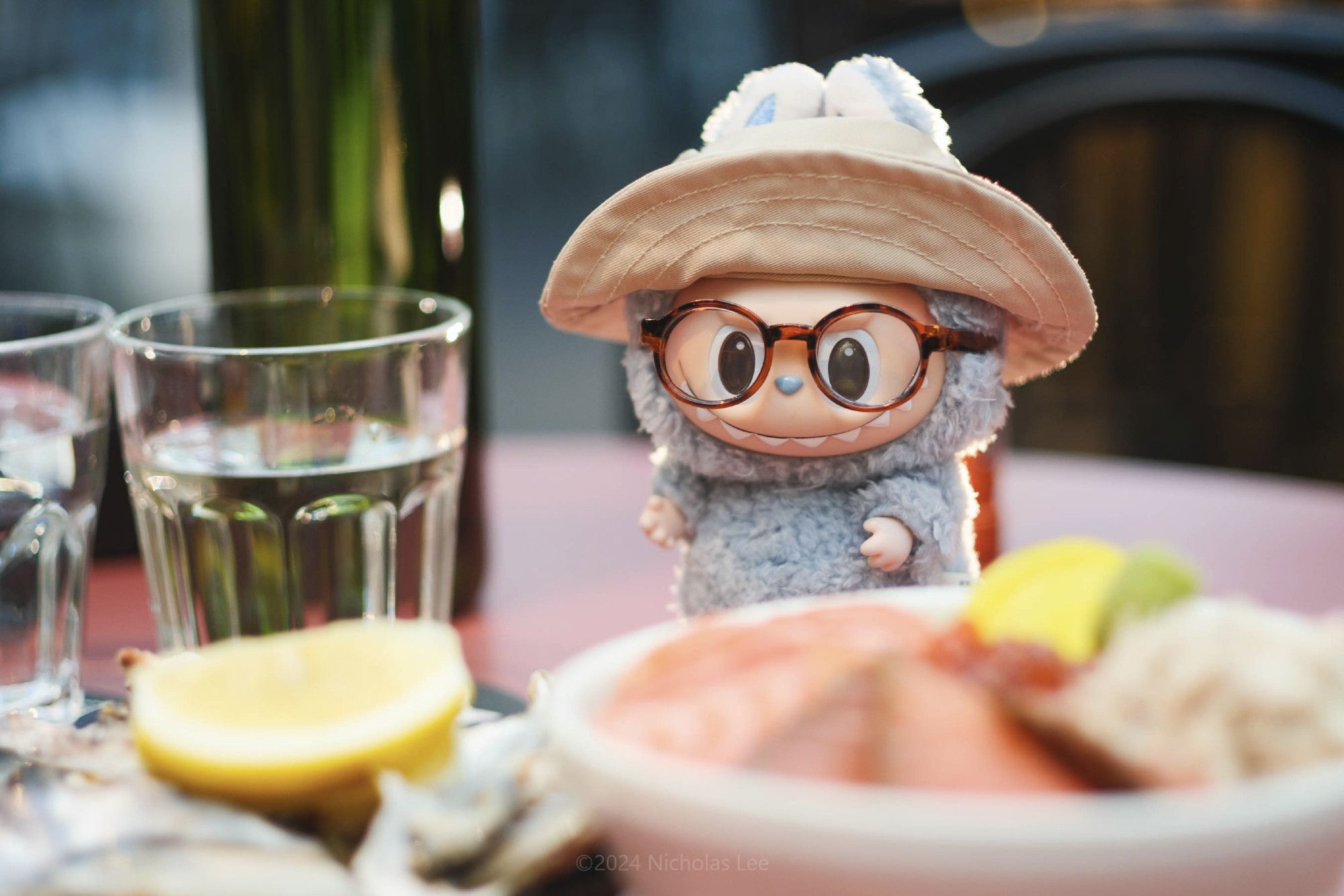 A photo of a Labubu toy wearing a sun hat and glasses standing amidst out of focus plates of food.