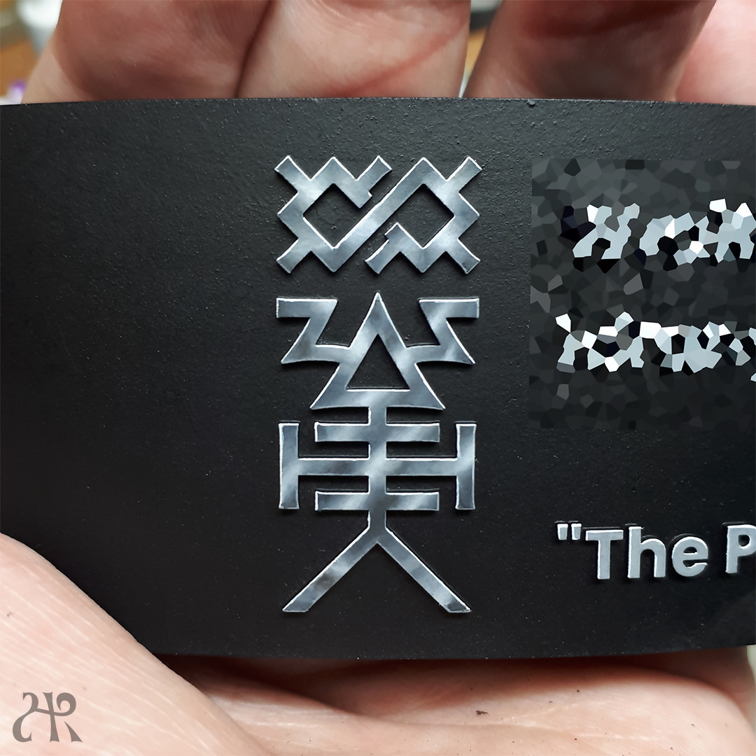 A hand holding a black plinth showing a non-metallic-silver rune that depicts a triangle with 2 "horns", 3 horizontal lines below it and two downwards and outwards pointing lines. Above it all is a pointy infinity-symbol. The text "The P" can be seen on the side.