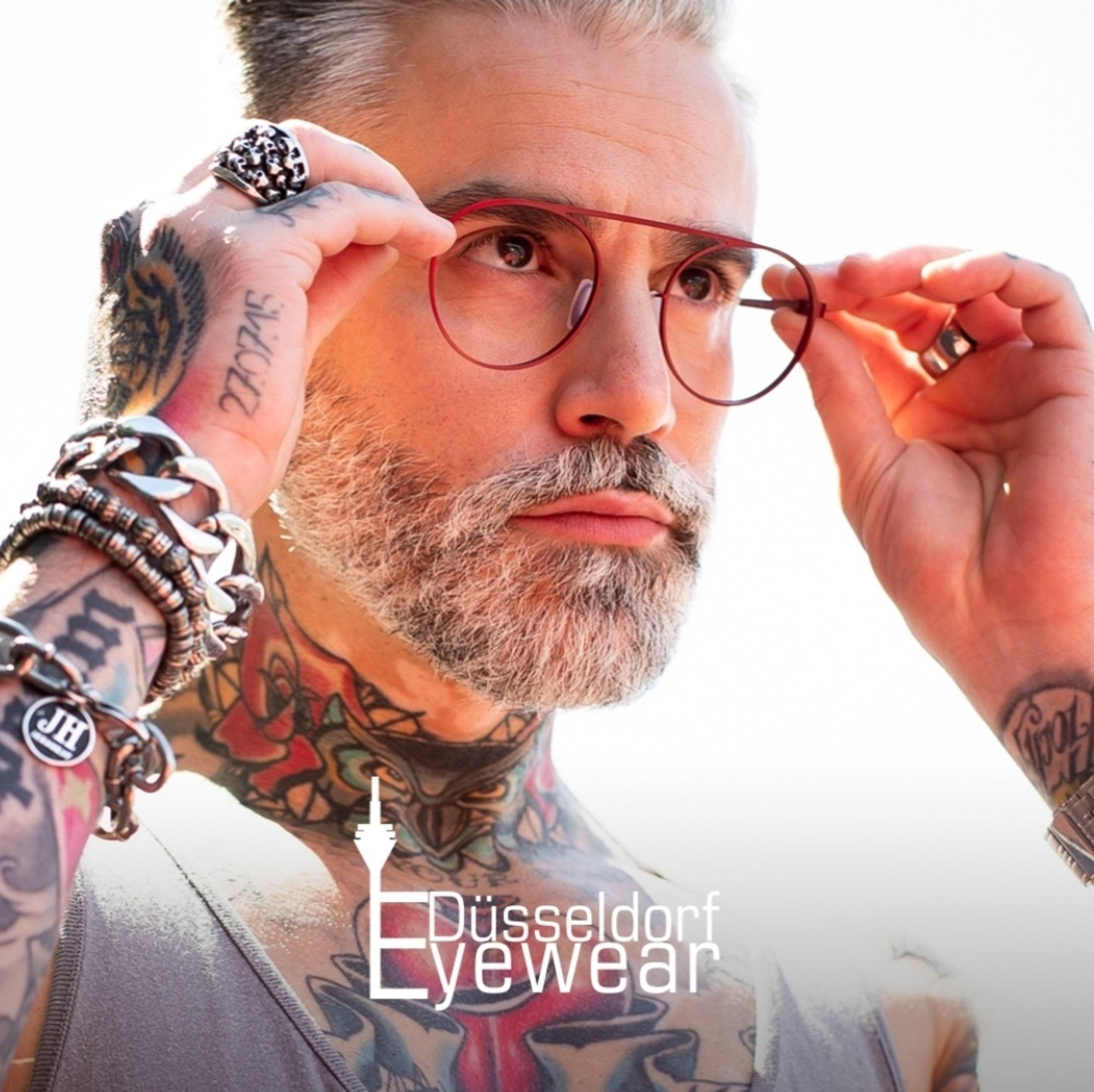 Werbebild von Düsseldorf Eyewear aus 2019, eine maskuline Person setzt sich eine rote Brille aus dünnem, matten Metall auf und blickt in die Ferne, sie hat viele Tattoos, grauen Bart und graue Kurzhaarfrisur und trägt ein Tanktop und Silberschmuck an Fingern und Handgelenken