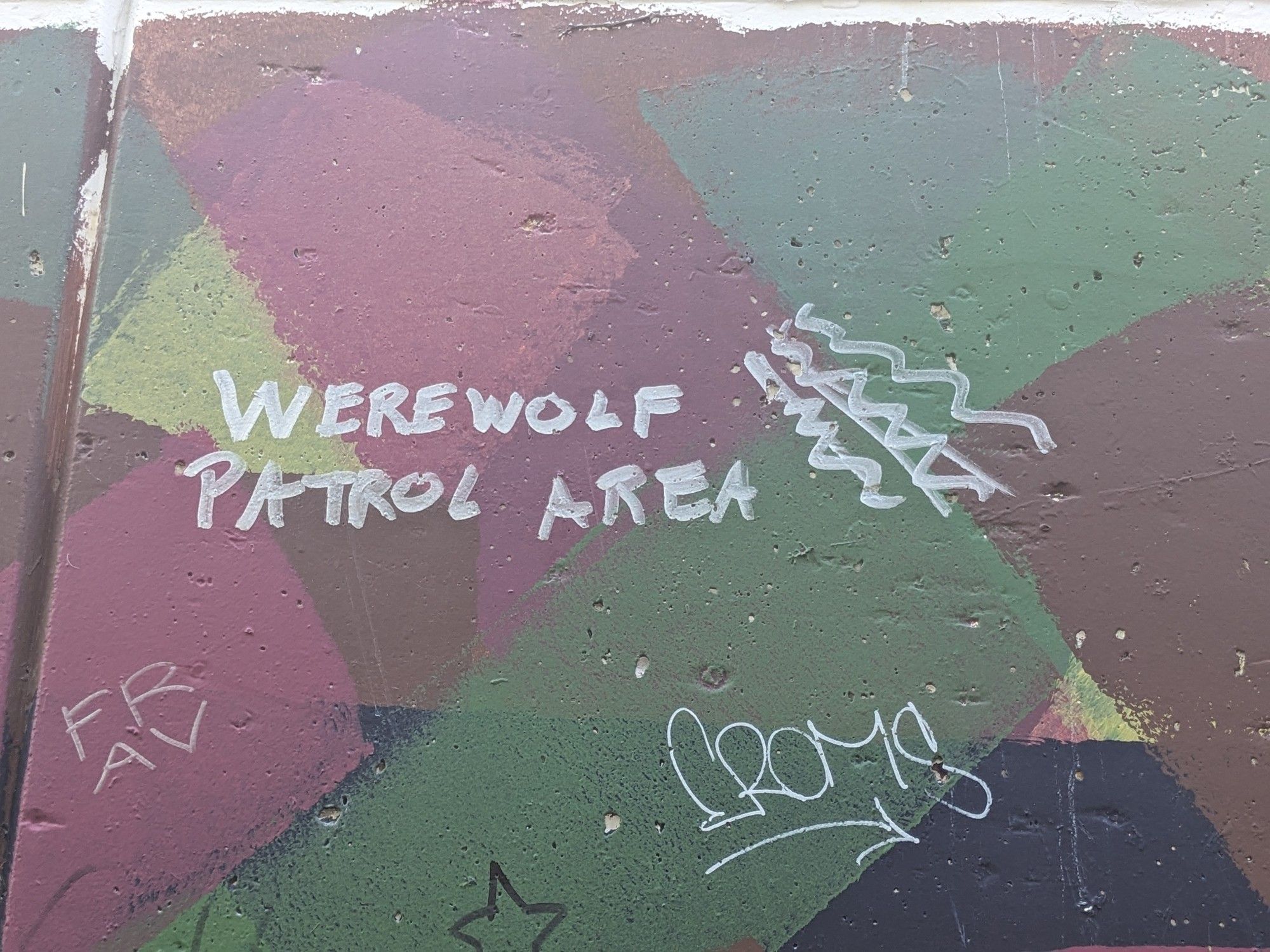 Part of a graffiti wall. In silver paint marker, the phrase "werewolf patrol area" is written with squiggly lines looking like stylized claw marks