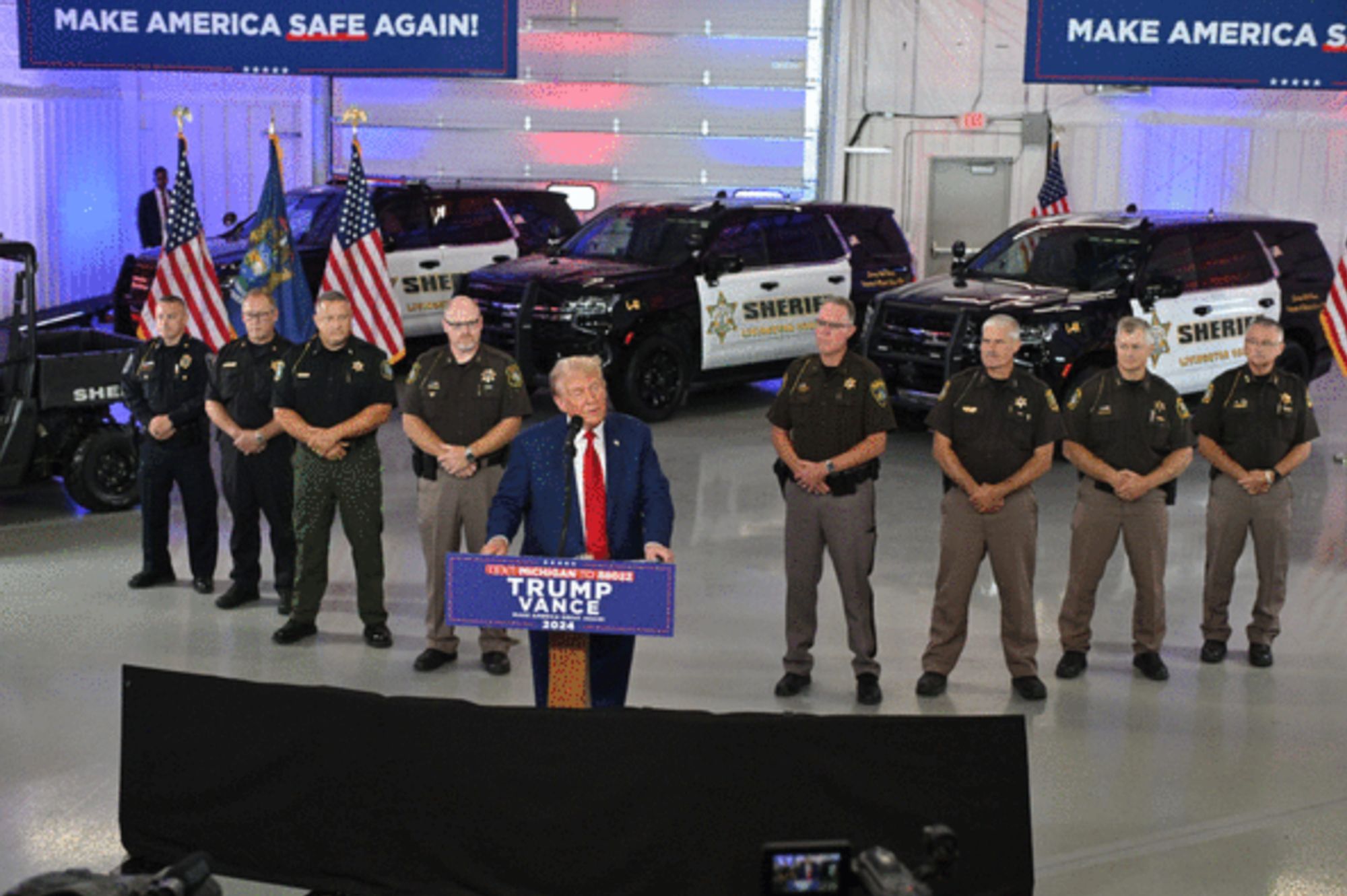 Trump at a podium in a parking garage with cop cars. He is flanked by flags and eight middle aged white sherifs in uniform who are standing with their hands folded like an honor guard. Signs above him say "Make America Safe Again!" (Crime in the US is at historic lows.) 

Source: Craig Mauger, The Detroit News on Aug 20, 2024

https://www.arcamax.com/politics/politicalnews/s-3377287