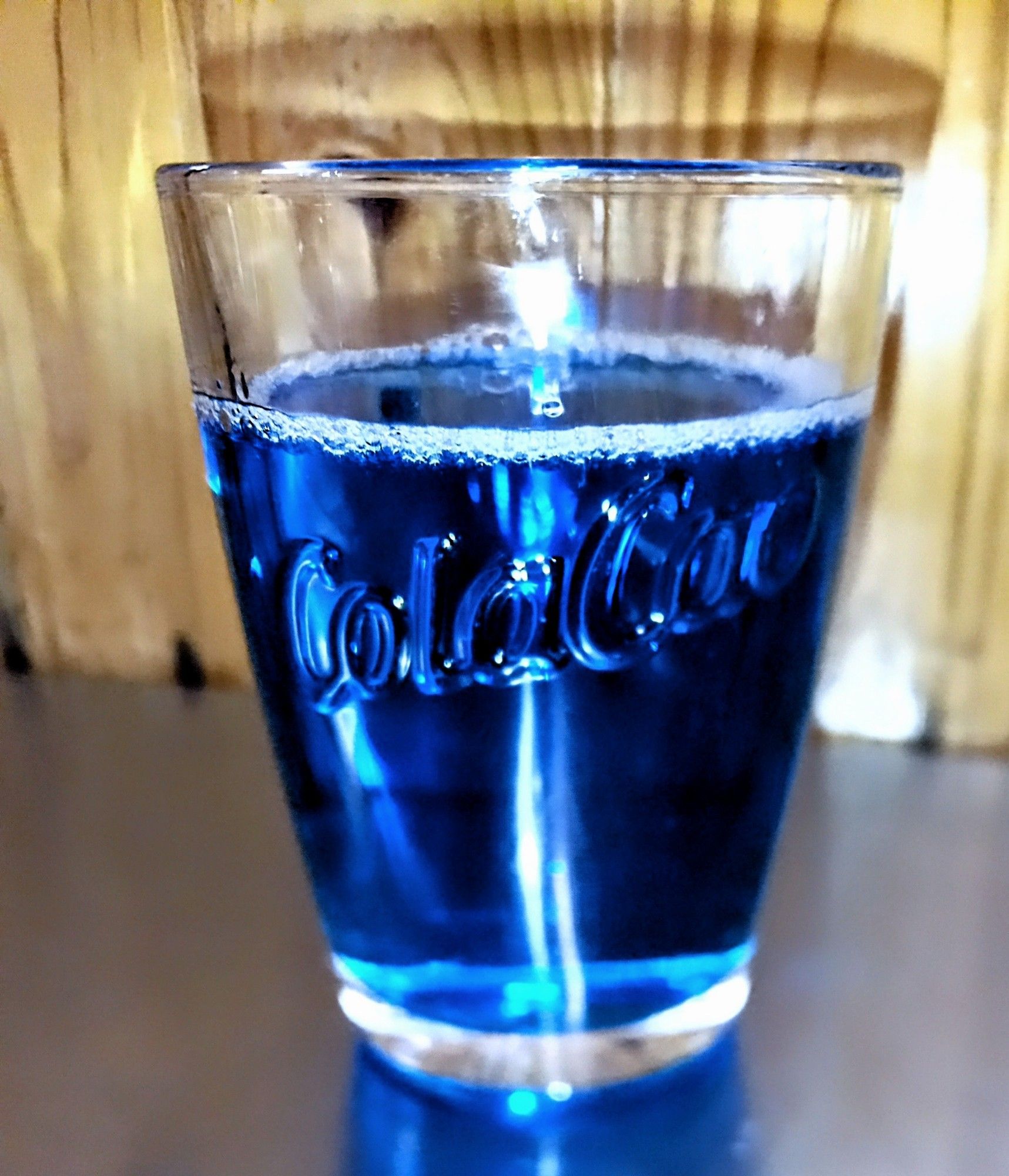 Un vaso con las letras ColaCao en relieve. Contiene un liquido azul brillante (abrillantador de lavavajillas industrial), con un aro de espumilla alrededor.