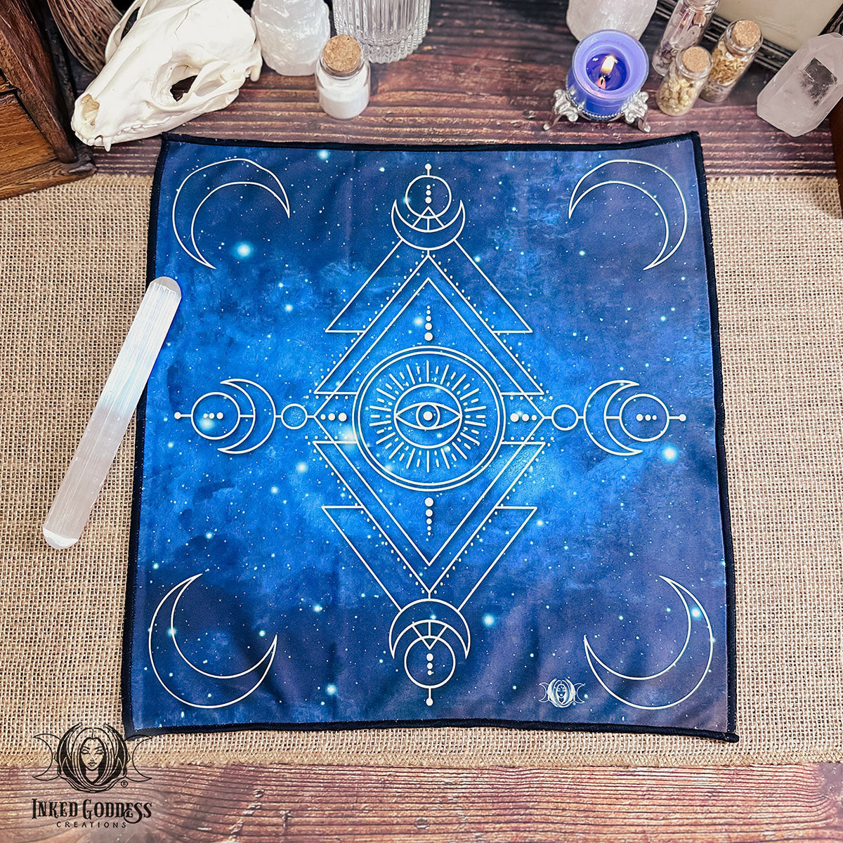 A Moon Magick Altar Cloth is spread on a burlap covered table surface with a selenite wand. The background is a watercolor space illustration with nebula clouds and stars. The other imagery includes a line drawing of mystical symbols like crescent moons and an evil eye talisman. This is an exclusive product from Inked Goddess Creations.