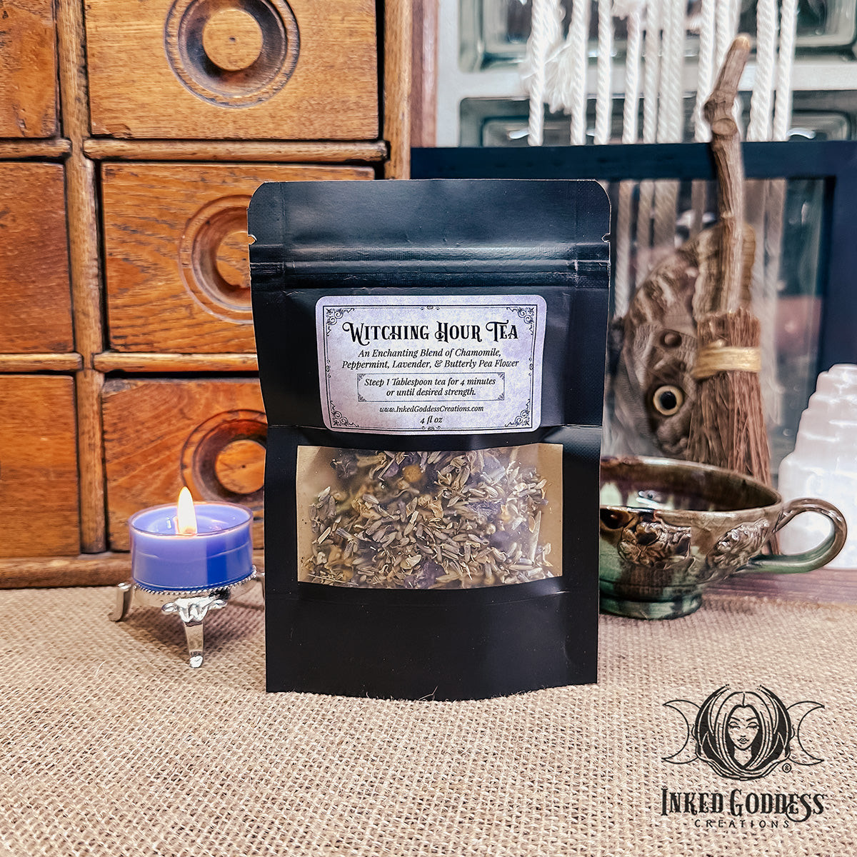 A pouch of loose leaf tea stands in front of a glazed earthenware teacup on a burlap-covered surface. The pouch has a clear window on the front with a label that features the name of the tea, "Witching Hour Tea," ingredients, and preparation instructions. This is an exclusive product from Inked Goddess Creations.