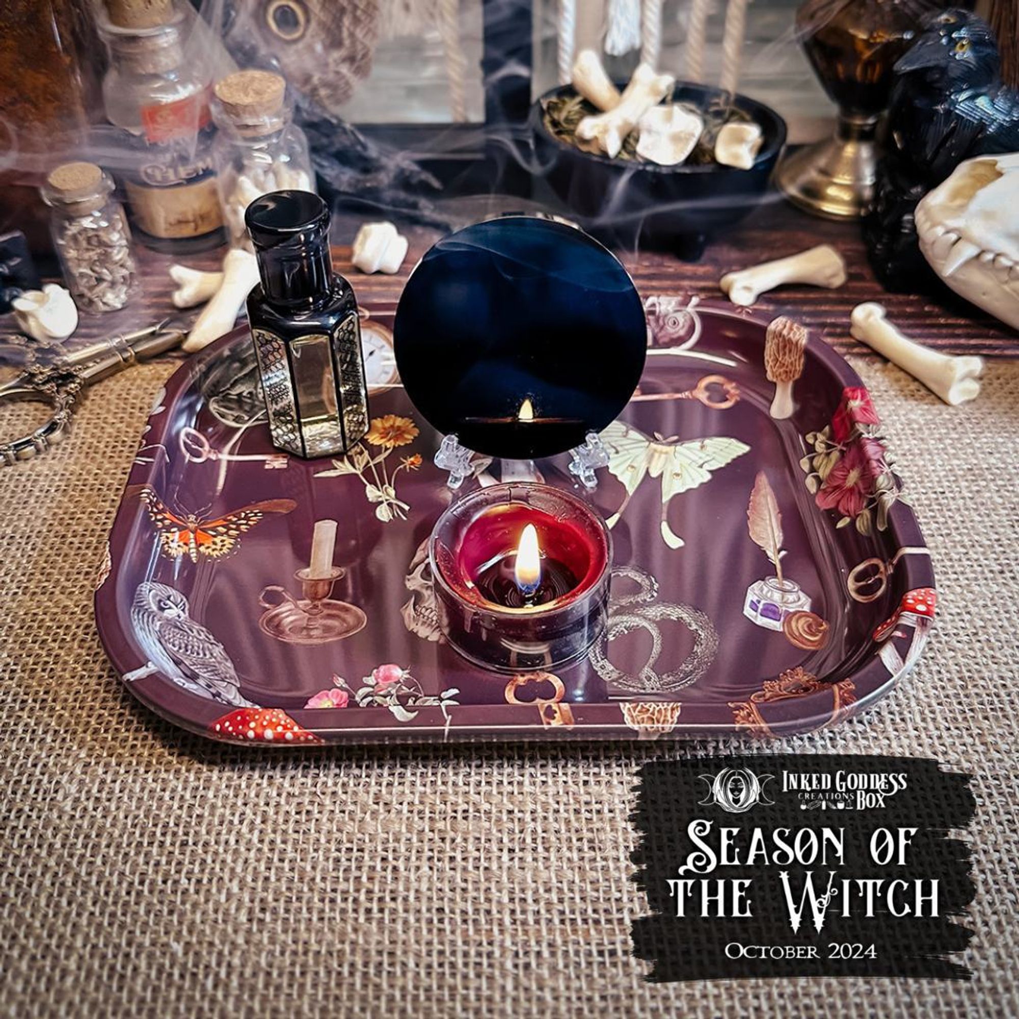 Three items are arrayed on a burlap-covered surface. The first is a smoked black glass scrying mirror placed behind a burning tealight candle. The second is a glass vial with yellowish liquid inside and patterns in black around the vial. They are all on a third item, a small tray covered in illustrations of witchy imagery. These are just three of the items in the exclusive October 2024 "Season of the Witch" Inked Goddess Creations Box.