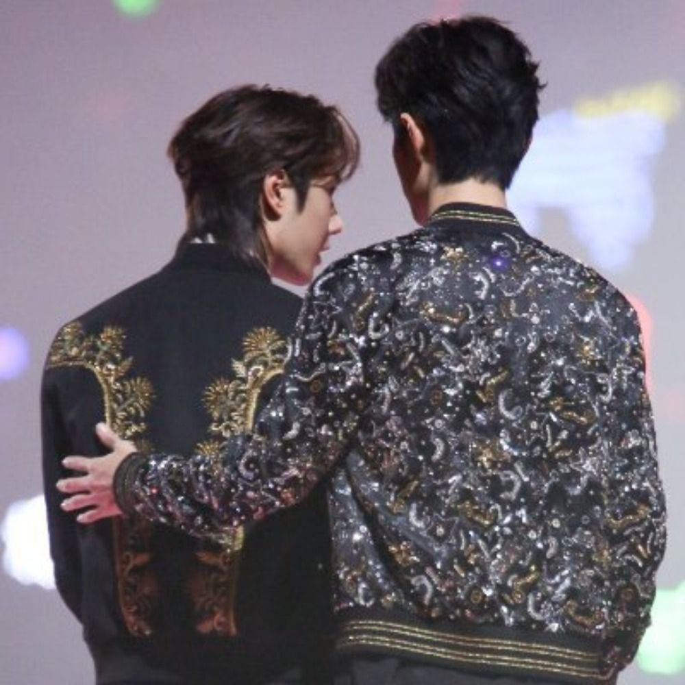 Wang Yibo (left) and Xiao Zhan (right)at the 2019 Tencent Starlight Awards. XZ places his left hand on Yibo's back. XZ is wearing a black, silver and gold sparkling jacket with moon and stars on it. Yibo is wearing a black jacket with two gold trims, on each side, going from his shoulder to the center bottom of his back.