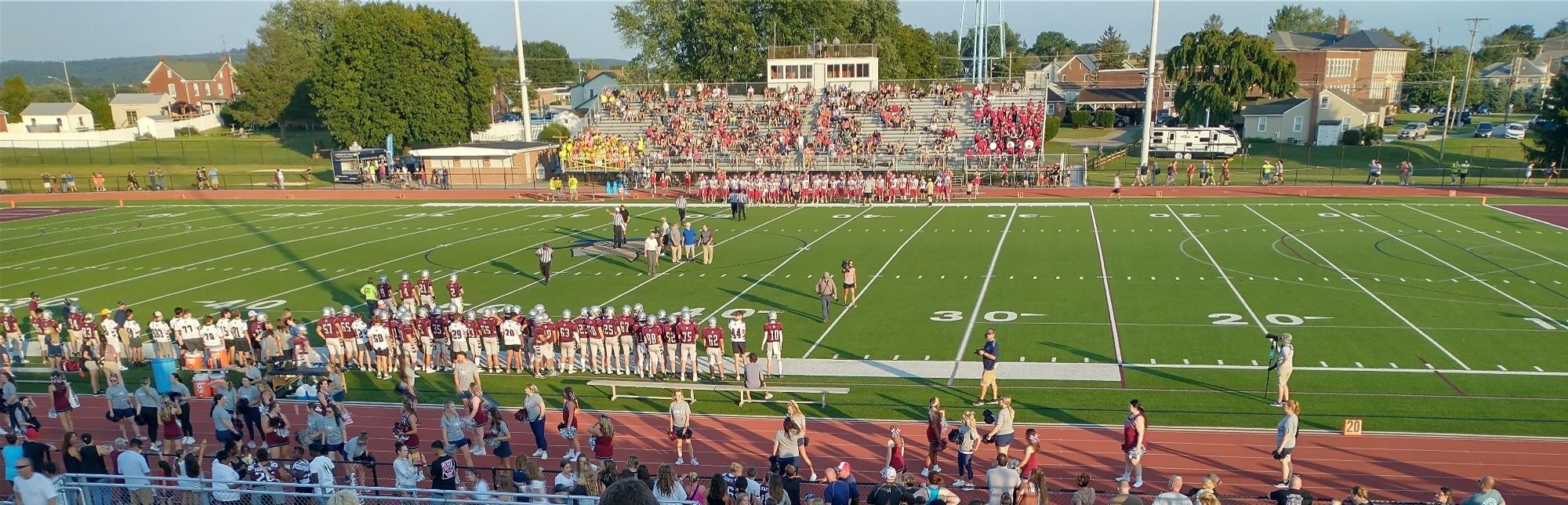 A sunlit green football field holds players in red and maroon while people fill the terra cotta colored track and the gray metallic stands across the field.