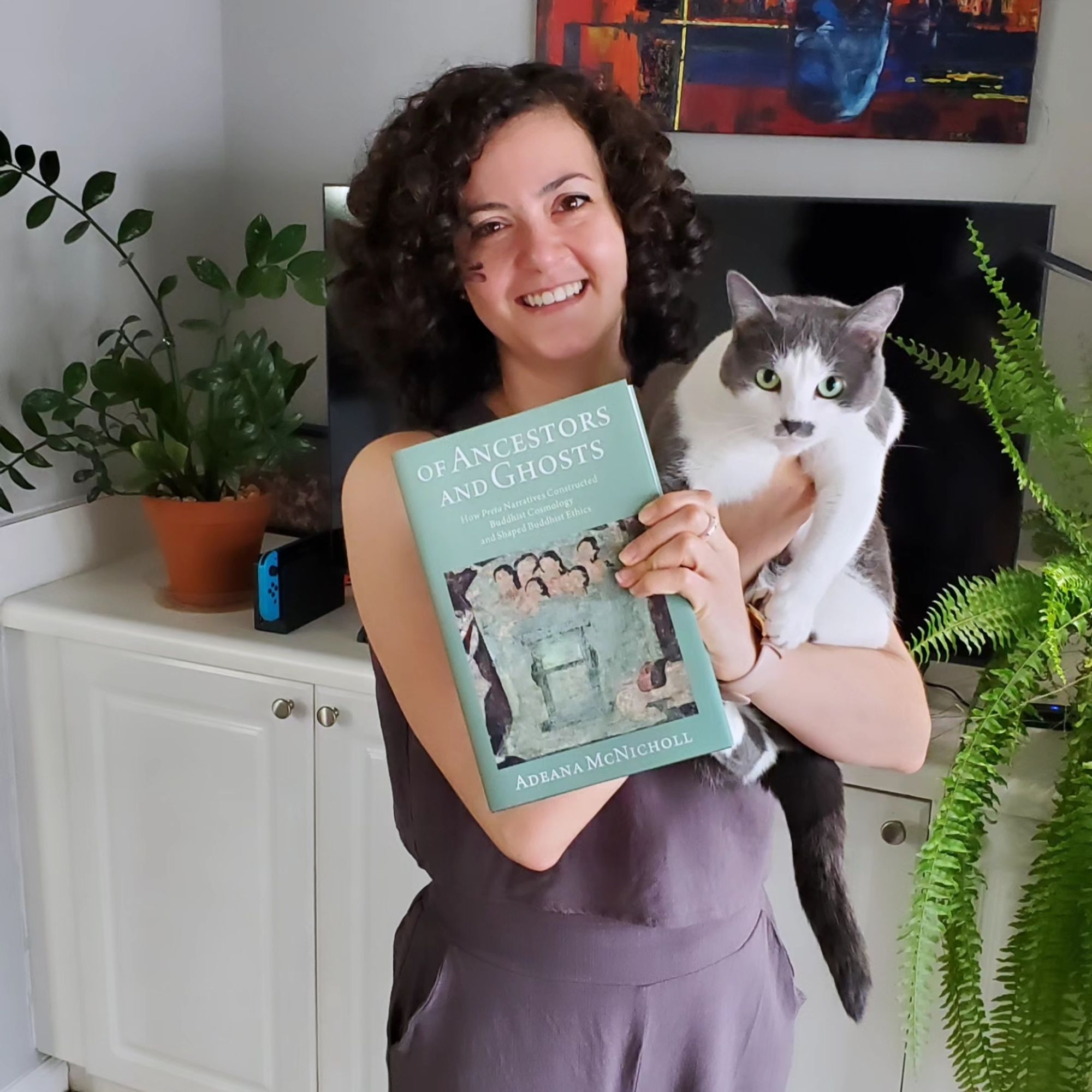 A woman holding a book in one hand and a cat with a mustache in the other. The book is titled Of Ancestors and Ghosts