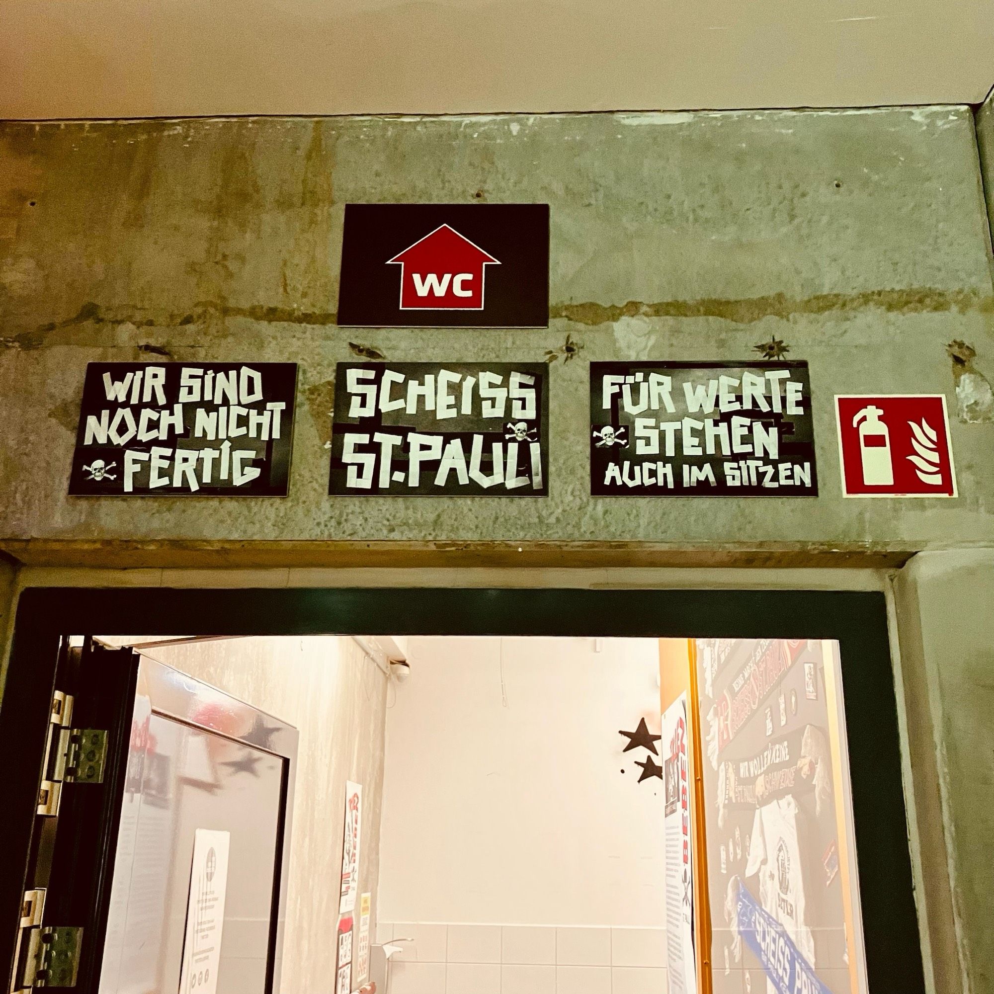 Über dem Gang zum WC in der Weinbar am Millerntor-Stadion hängen drei Schilder:

WIR SIND 
NOCH NICHT 
FERTIG

SCHEISS 
ST.PAULI

FÜR WERTE STEHEN 
AUCH IM SITZEN

http://www.keinweindenfaschisten.de/weinbar/