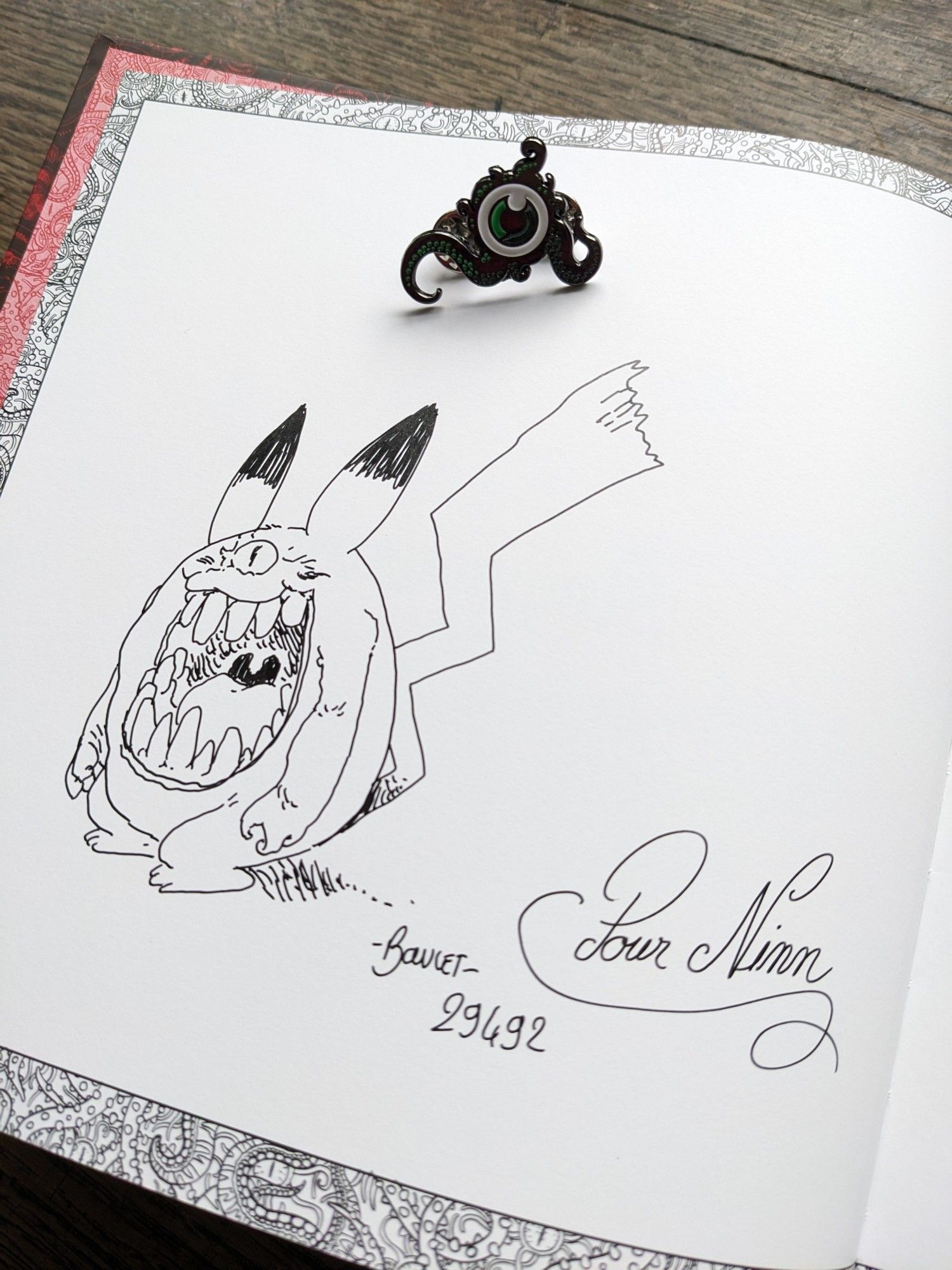 The same book, open to an autograph by the author, with a pen and ink drawing of a monstrous, one-eyed Pikachu. There is also a small metal brooch depicting an green eye surrounded by tentacles.