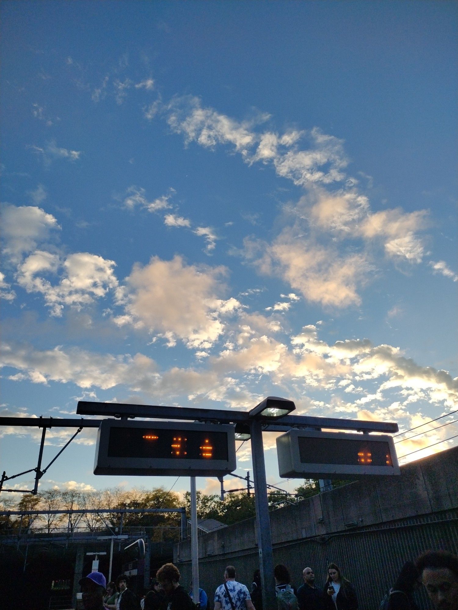 The golden hour at a train station. Shimmering clouds in a blue sky, trees beginning to change color on an overpass.