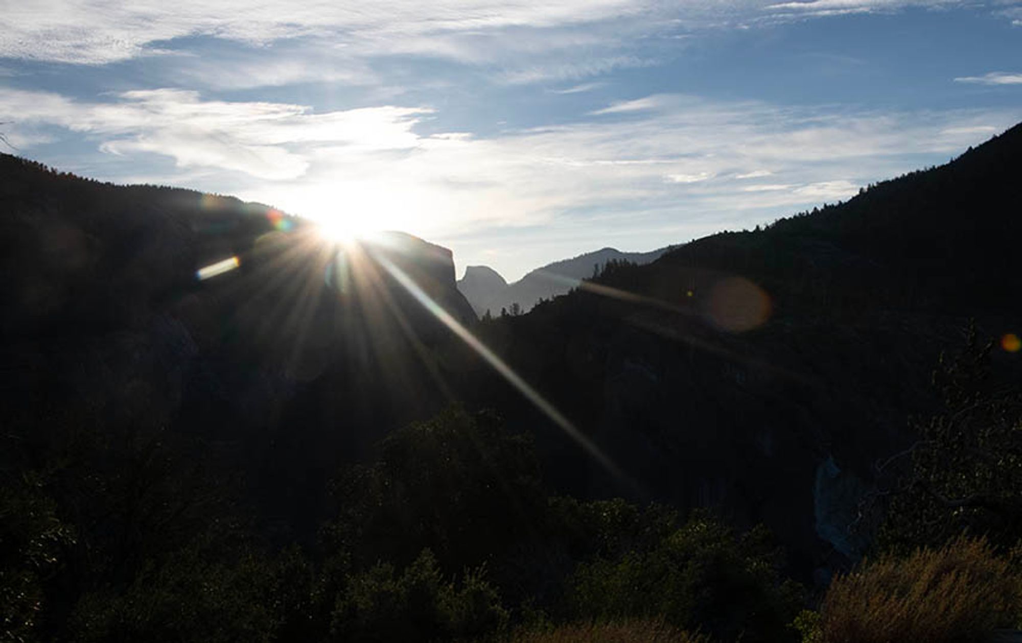 The sun rises in a starburst over El Capitan. Half Dome can be seen in the background. El Capitan and Half Dome are in silhouette.