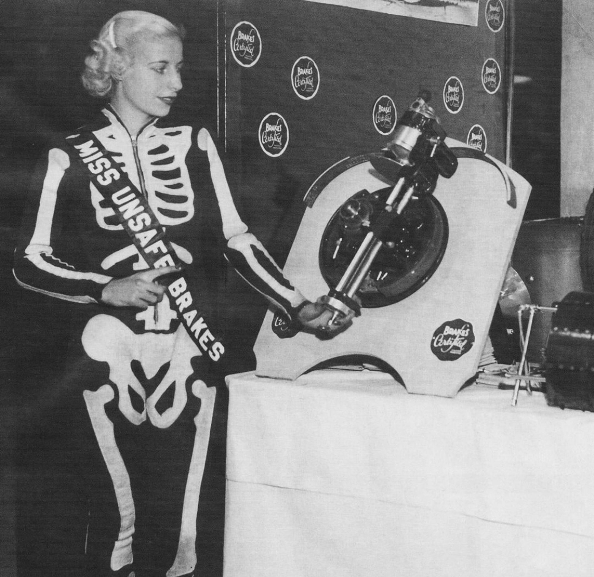 An old photo of a woman at a trade show in a skeleton print onesie and a sash that says “miss unsafe brakes” looking at some exhibit about brakes