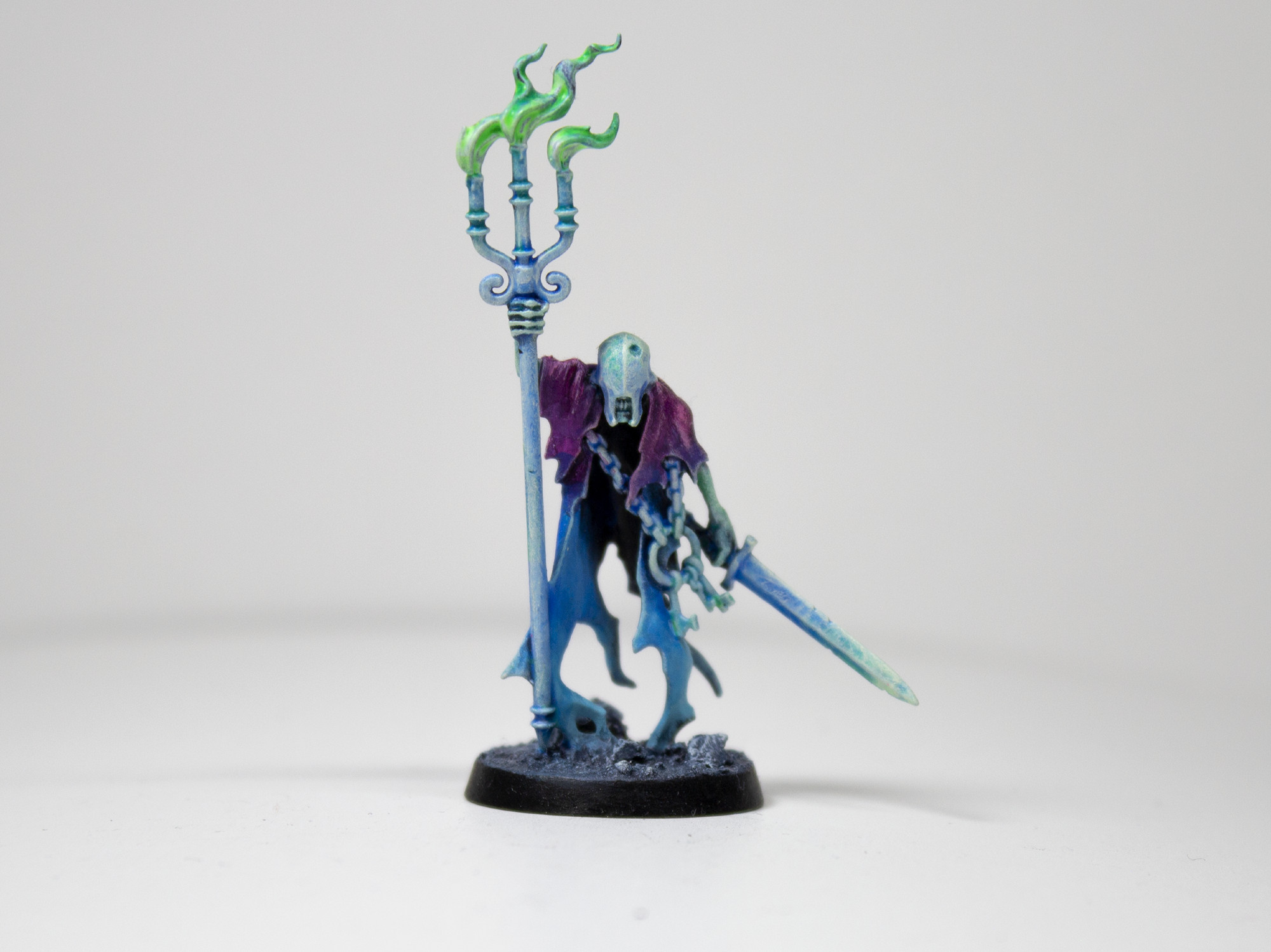 Painted miniature of a cloaked, ghastly figure raising a staff-like candelabra. It is painted in a gradient from a bright purple to a lighter blue, and the flames of the candelabra are an unnatural green