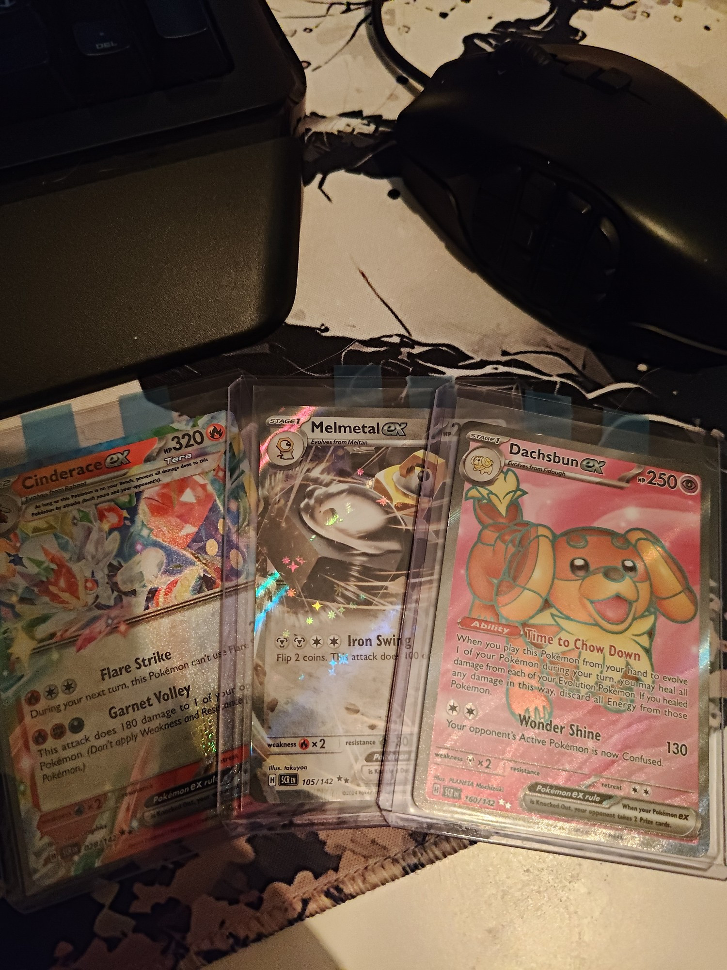 3 pokemon cards, Cinderace EX, Melmetal EX and Daschbun EX, sleeved in top loaders next to a mouse and keyboard.