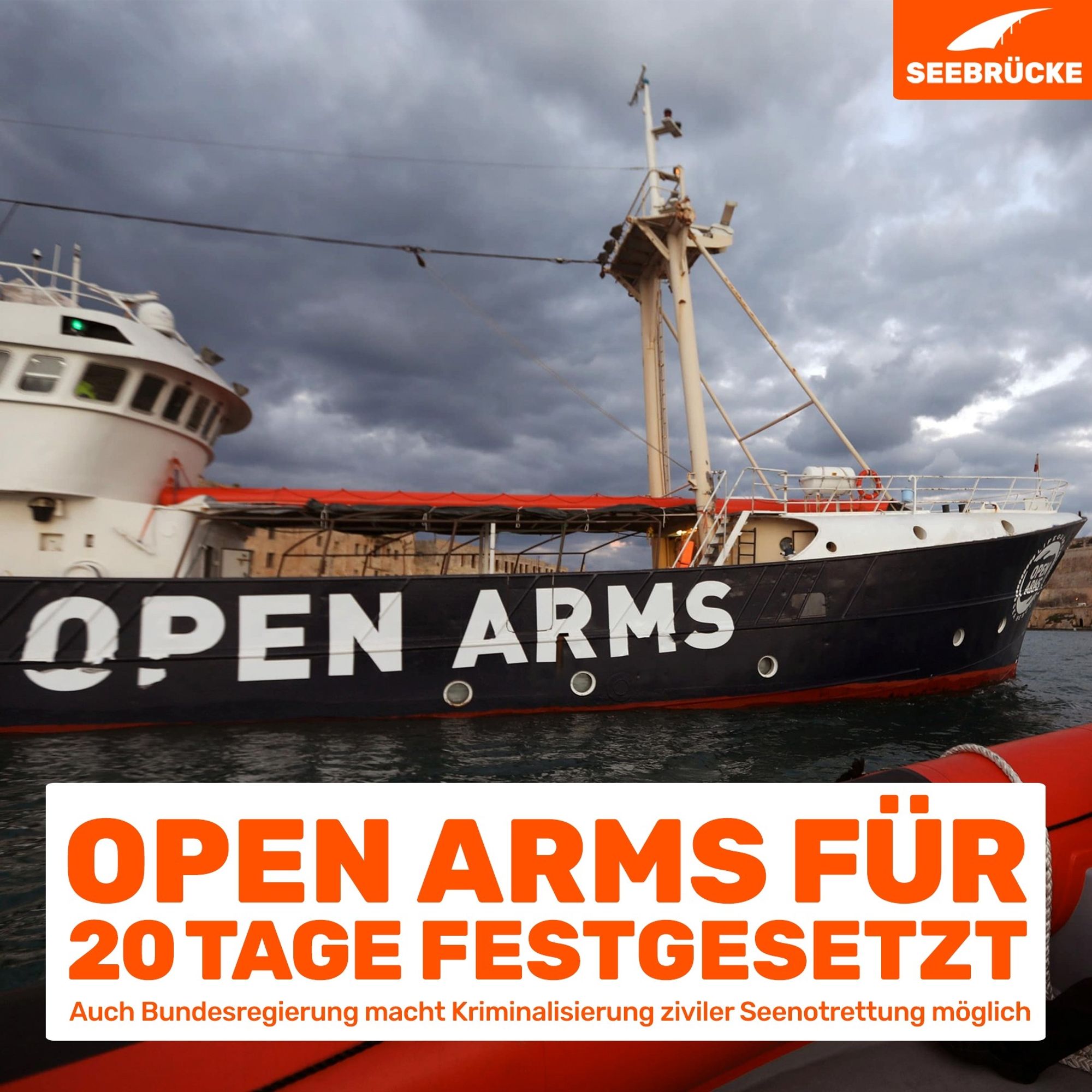 Foto der Open Arms von einem Beiboot aus. In einem weißen Kasten steht "Open Arms für 20 Tage festgesetzt. Auch Bundesregierung macht Kriminalisierung ziviler Seenotrettung möglich" in der rechten oberen Ecke ist das Seebrücke-Logo in weiß auf orange.