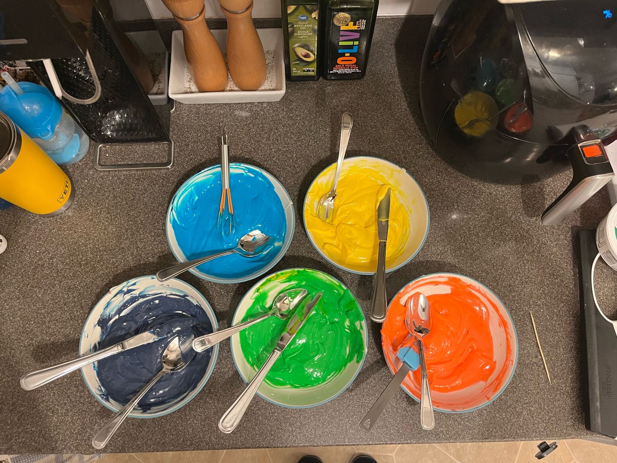 Five small bowls of cake batter sit on a kitchen counter, photographed from above. Each bowl has been stewed a different colour: red, yellow, green, blue, & purple.