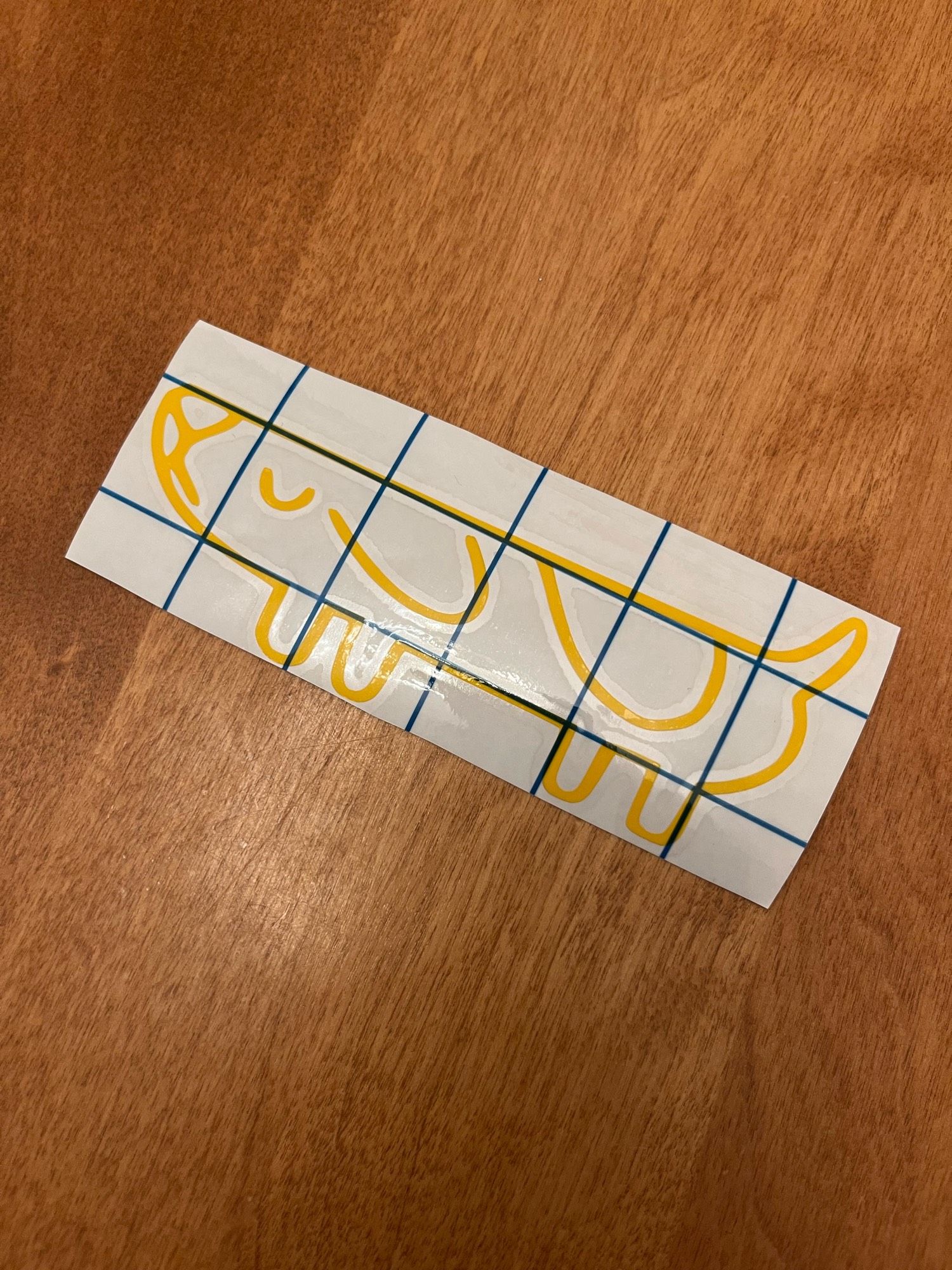 A sticker of a yellow outline of a “long dog” from Bluey, laid on a wooden tabletop. The sticker is on white backing and has a black grid over the top.