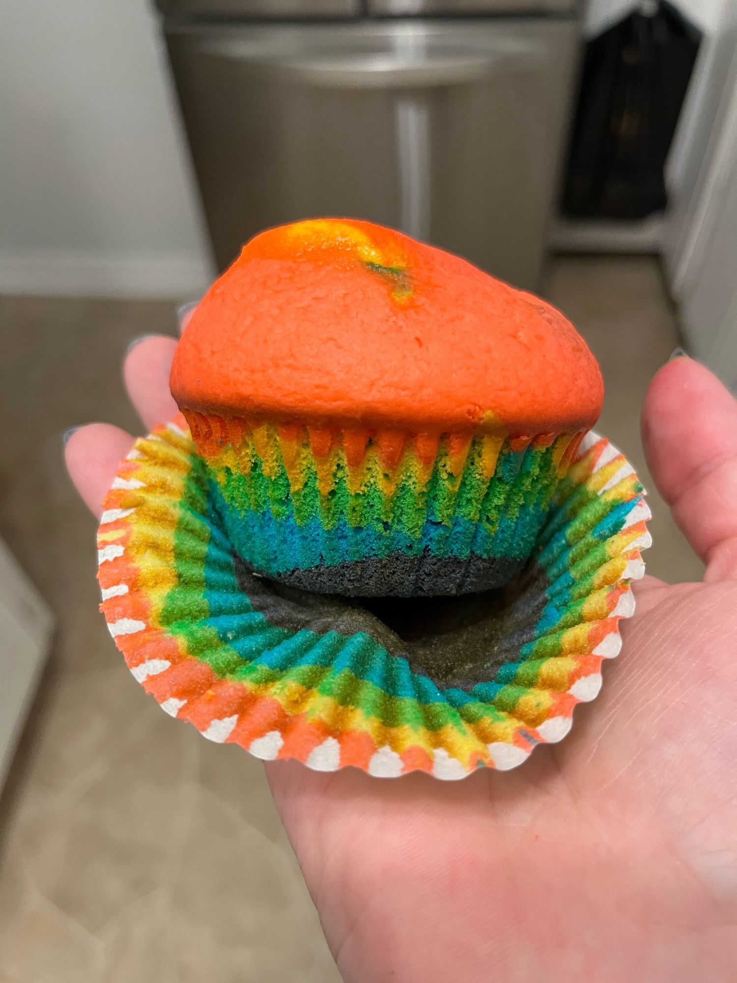 A cupcake, peeled out of its wrapper, she’s on a white woman’s hand. It has five layers of colour, in a rainbow, with red at the top and purple at the base.