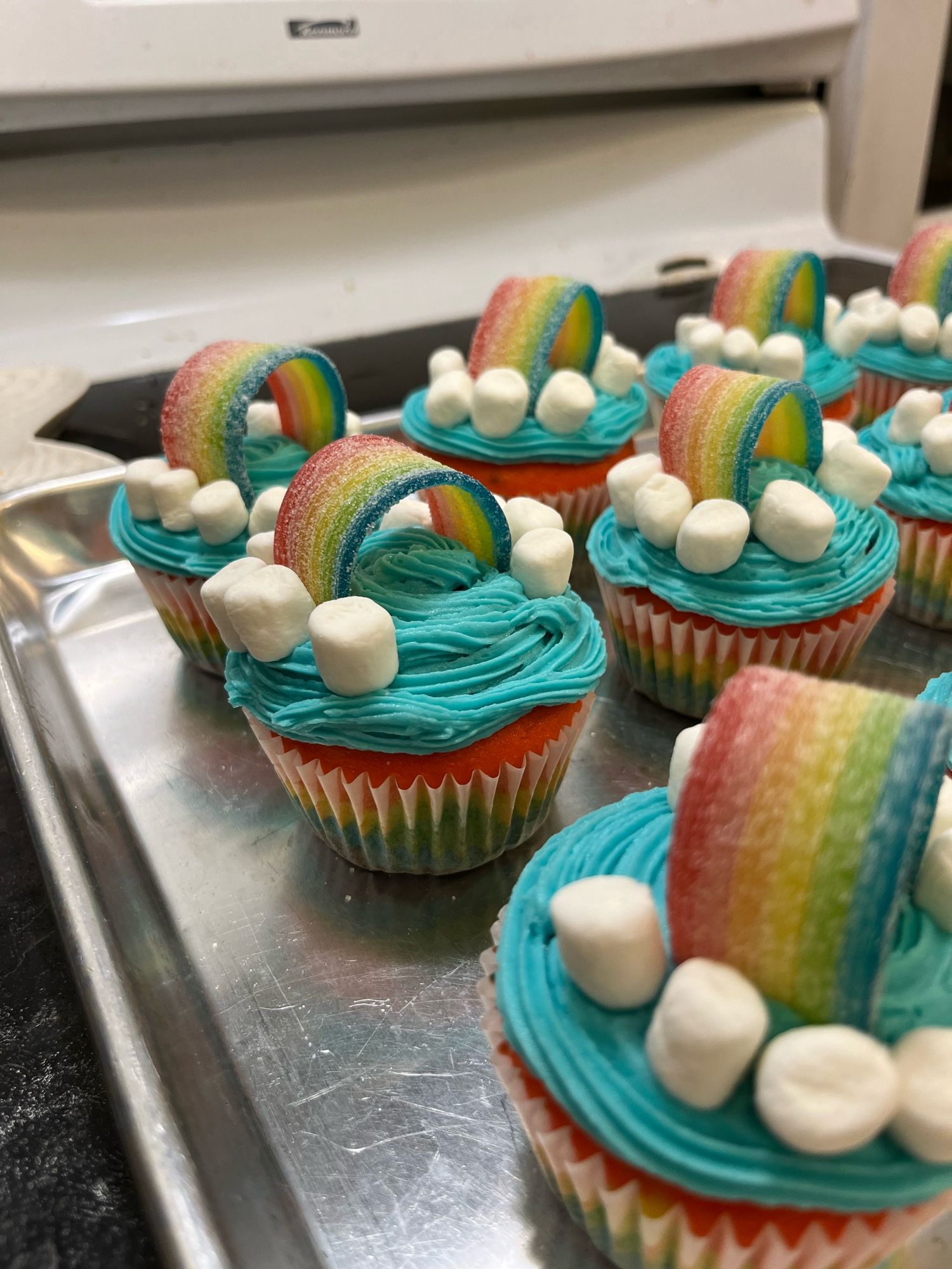 Several decorated cupcakes on a shiny silver baking pan. They are rainbow striped in the bottom, with blue icing and rainbow sour belts on top, with mini marshmallows for clouds.