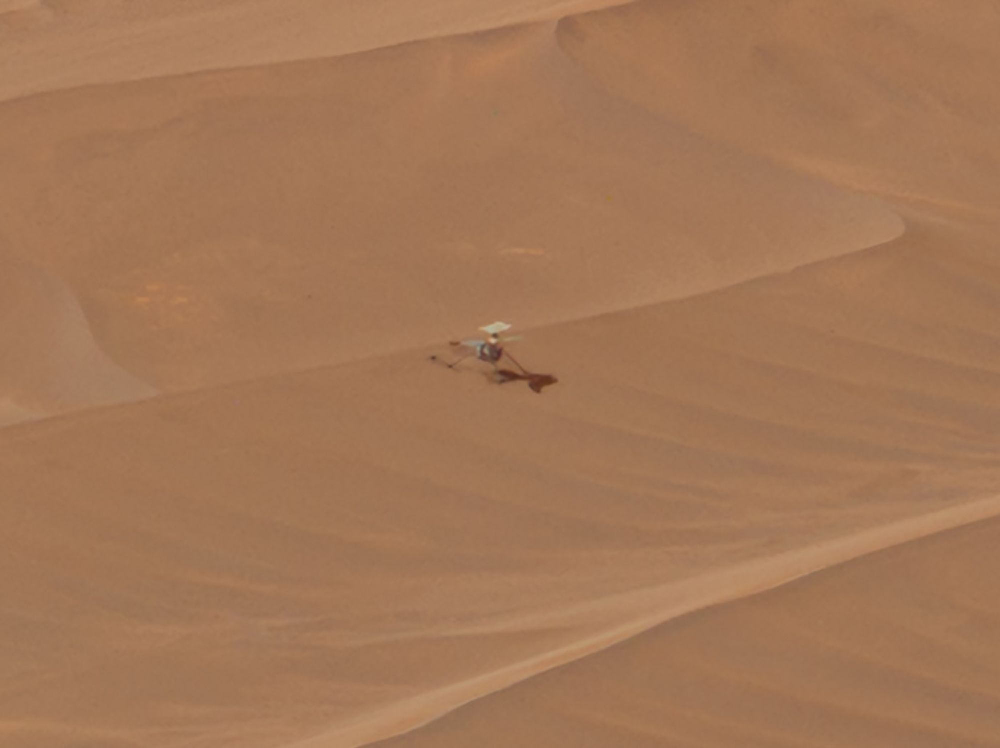 the Mars Helicopter standing on the slope of a sand ripple. two small dark marks can be seen in the sand next to her, these were presumably made by the impact of the rotor blades.