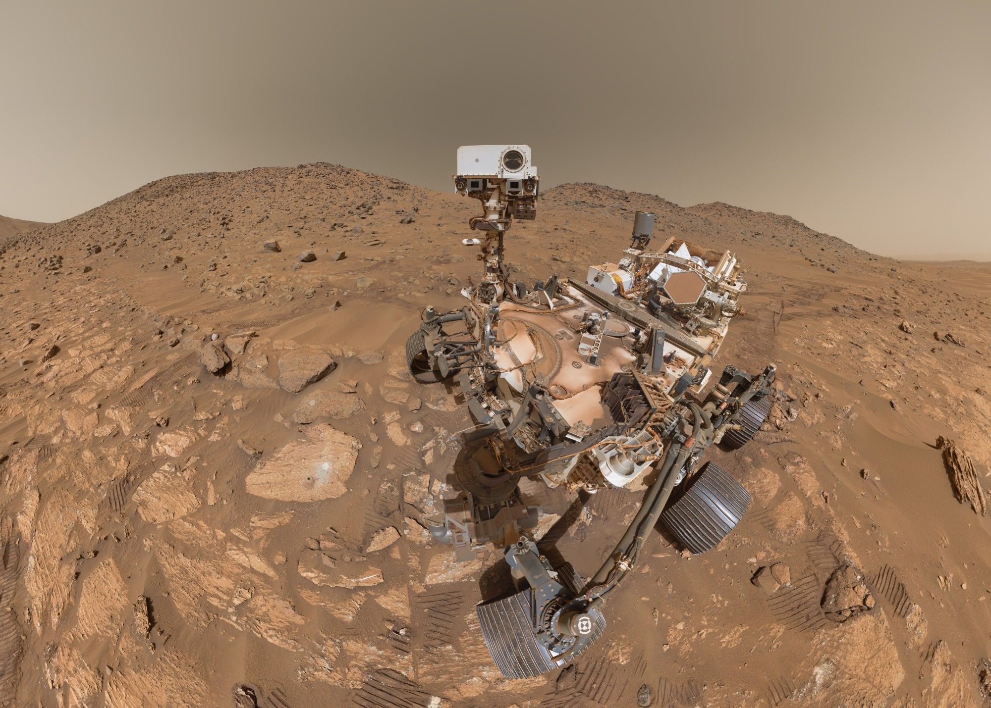 Self portrait of the dusty Perseverance rover standing in a field of reddish rocks. in front of her on the ground are two circular rock abrasions and one borehole where she took her latest sample.