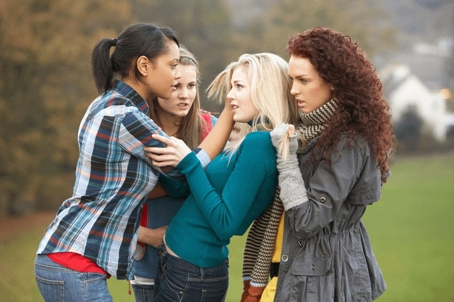 A stock photo of four women. A woman with curly auburn hair wearing a scarf, a woman with a black ponytail wearing a flannel shirt, and a woman wearing a Marty McFly-ass puffy vest are surrounding and bullying a woman with pale blonde hair wearing a turquoise shirt. Curly hair is pulling on a bit of blonde girl's hair in a fashion that looks almost playful, while ponytail rests a hand on the side of her face. The sexual energy is frankly, overwhelming. 