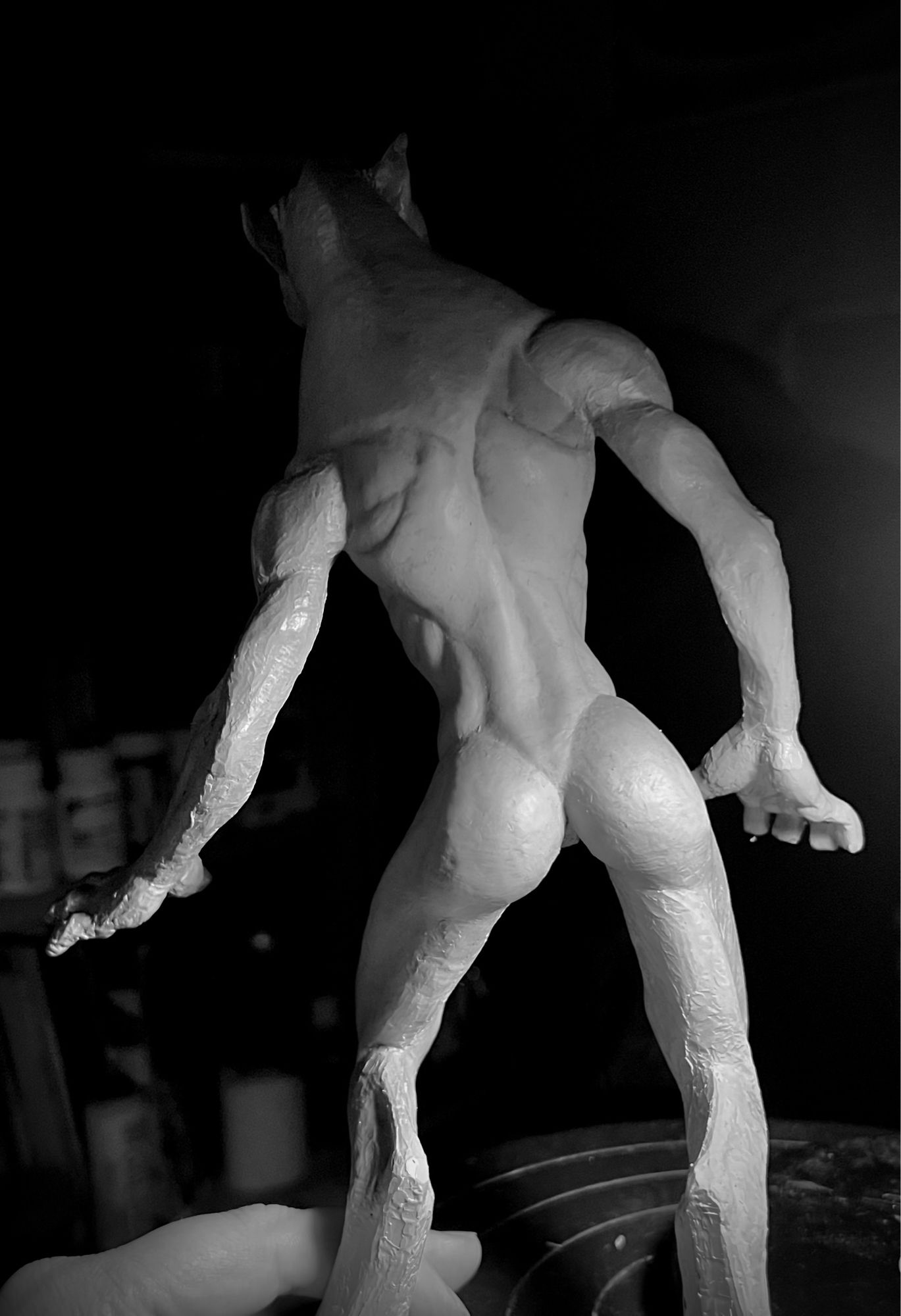 Dramatically lit photo of a werewolf sculpture viewed from the back. She is bipedal and has long, lithe limbs and a lean physique. Her arms end in appendages that are somewhere between paws and human hands. Her lower limbs are digitigrade, and terminate in large paws with dewclaws.