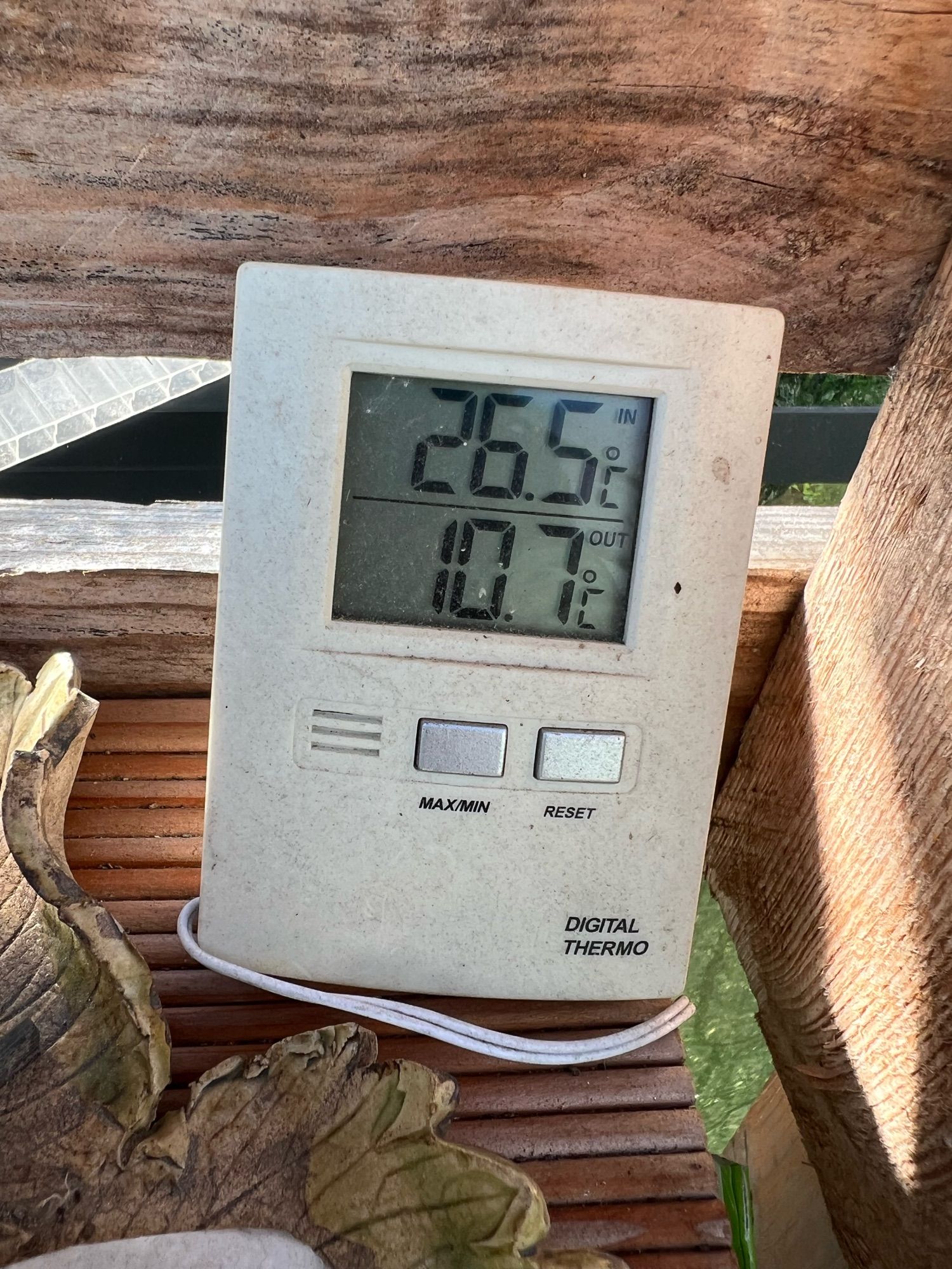 Ein weißes Digital Thermometer auf einem Holzregal. Innentemperatur 26,5°, Außentemperatur 10,7°