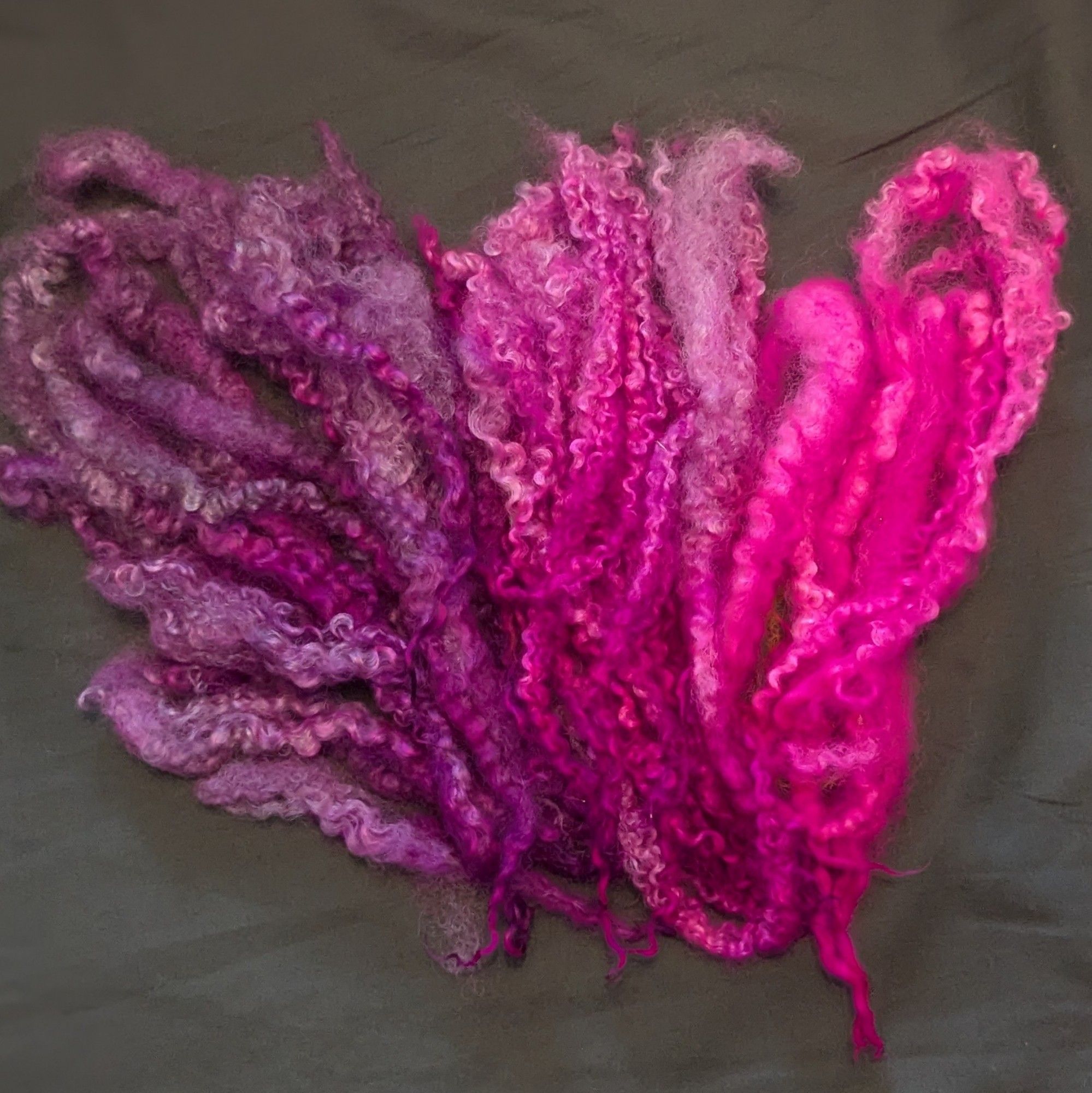 Hand dyed Masham locks on a grey background. The right is the brightest fluorescent pink, next to that slightly darker, and a beautiful black cherry on the left. All glow under blacklight to varying degrees.