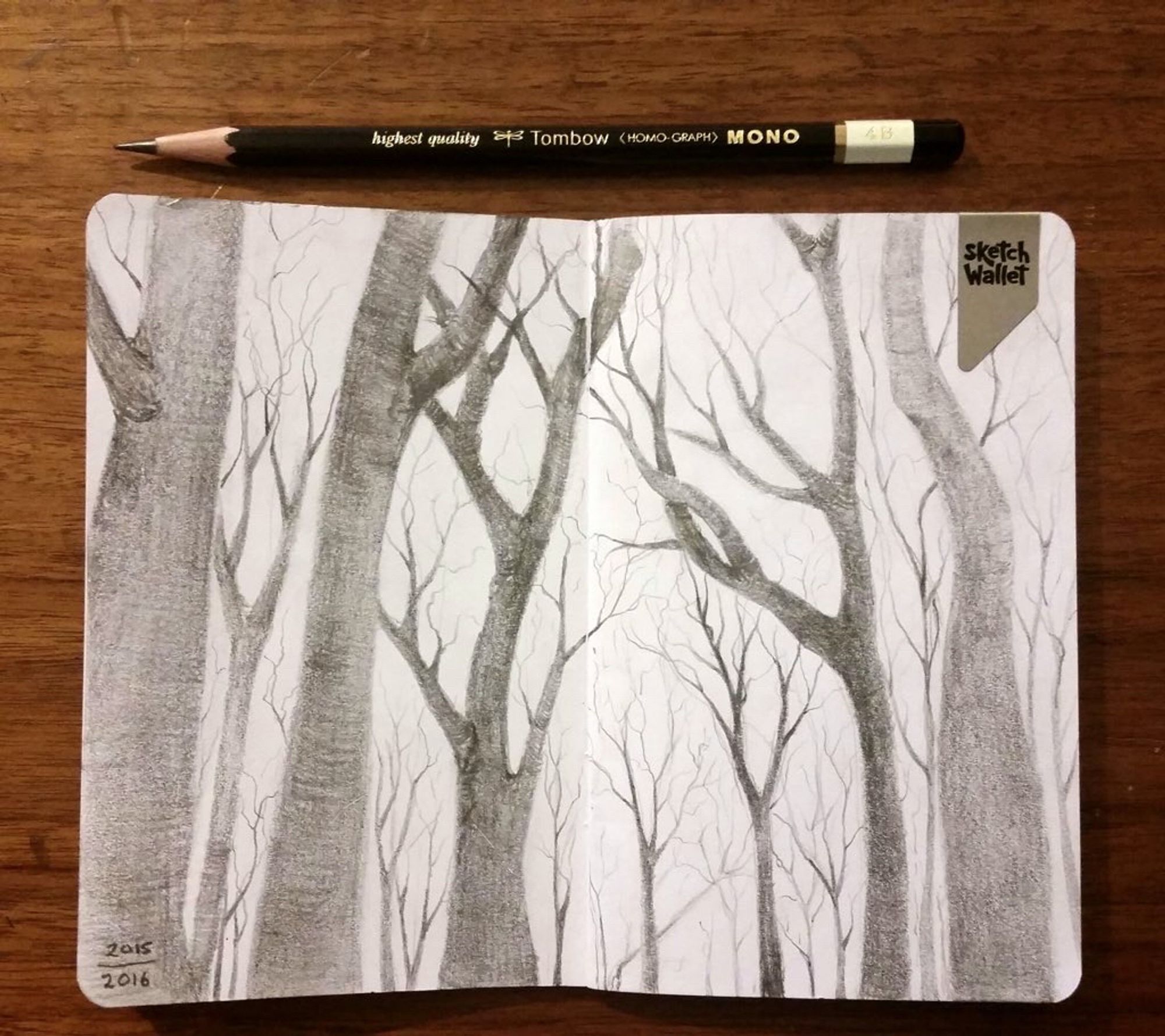 Sketchbook page, opened flat on a table. The spread features a forest in winter, drawn in pencil