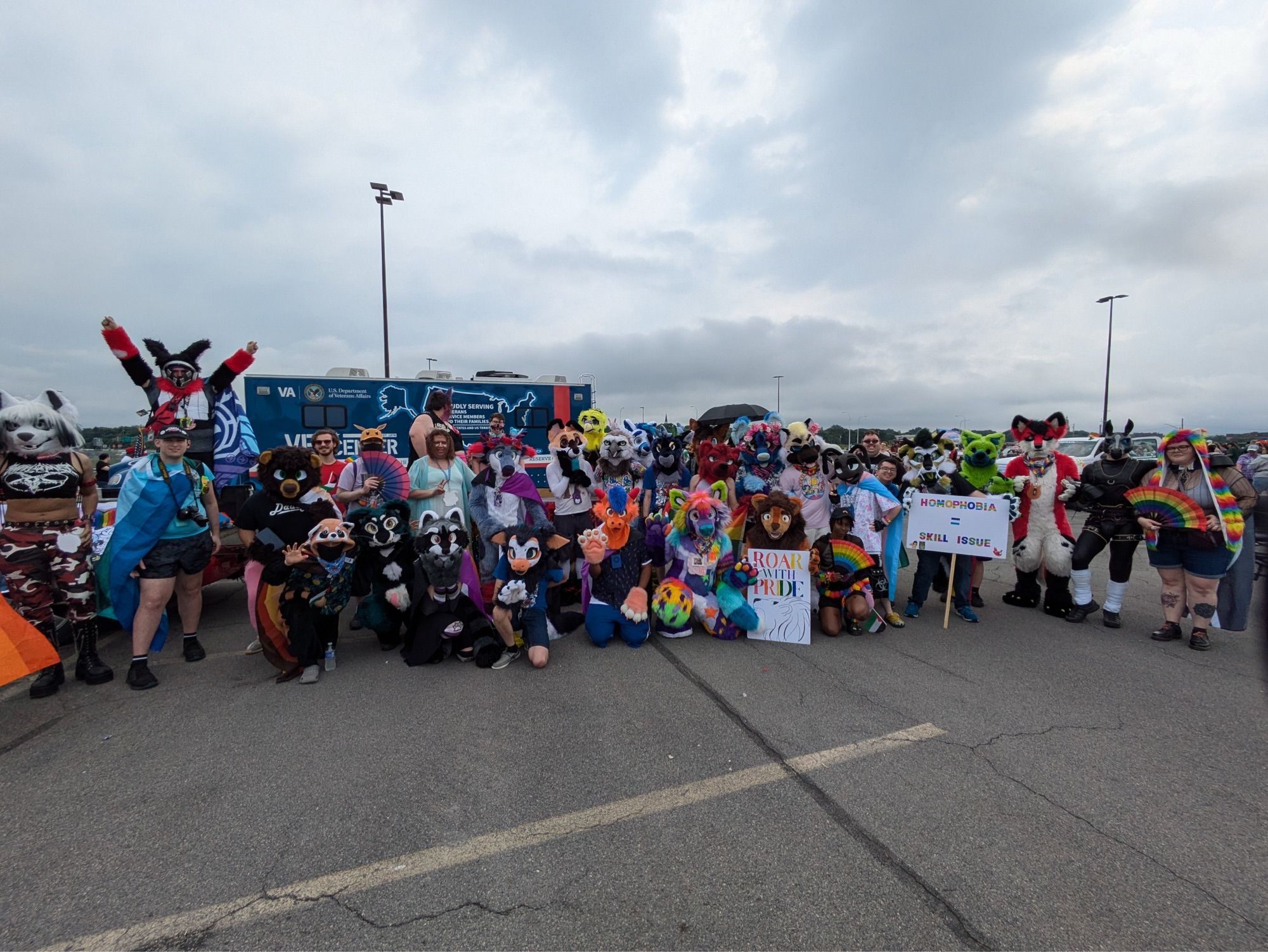 A groupshot of multiple fursuiters in front of a LGBTQ-themed parade float. Several of them are holding homemade signs.